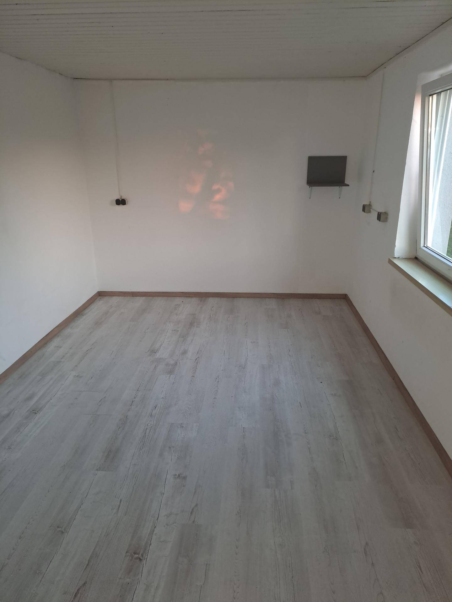 Leerer Raum mit hellem Laminatboden und einem Fenster, bereit zur individuellen Gestaltung.