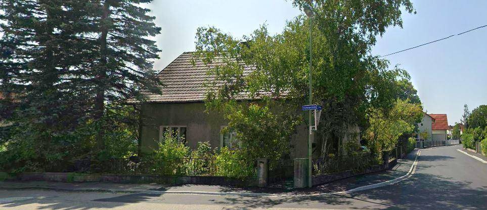 Eckansicht des Bestandsgebäudes mit Satteldach, umgeben von Bäumen und einer ruhigen Straße.