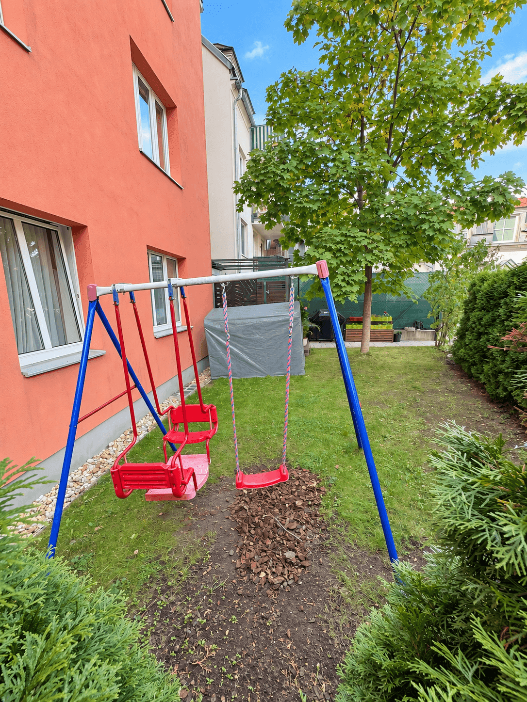 Garten mit Schaukelbereich und Rasenfläche neben dem Gebäude, umgeben von grünen Hecken.