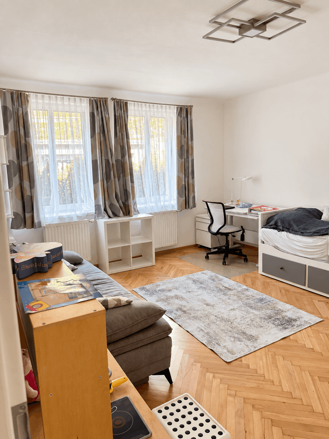 Freundliches Schlafzimmer mit Parkettboden, zwei Fenstern und einem praktischen Schreibtischbereich.