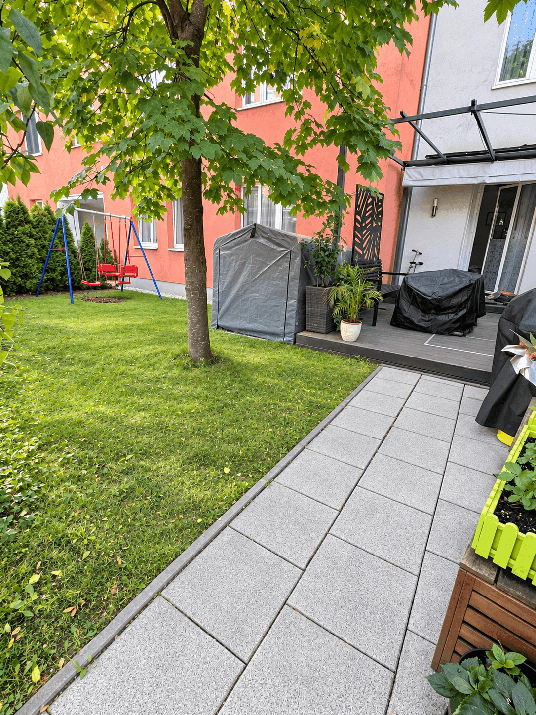 Gepflegter Garten mit Rasenfläche, Steinweg und Zugang zur Terrasse, umgeben von Bäumen.