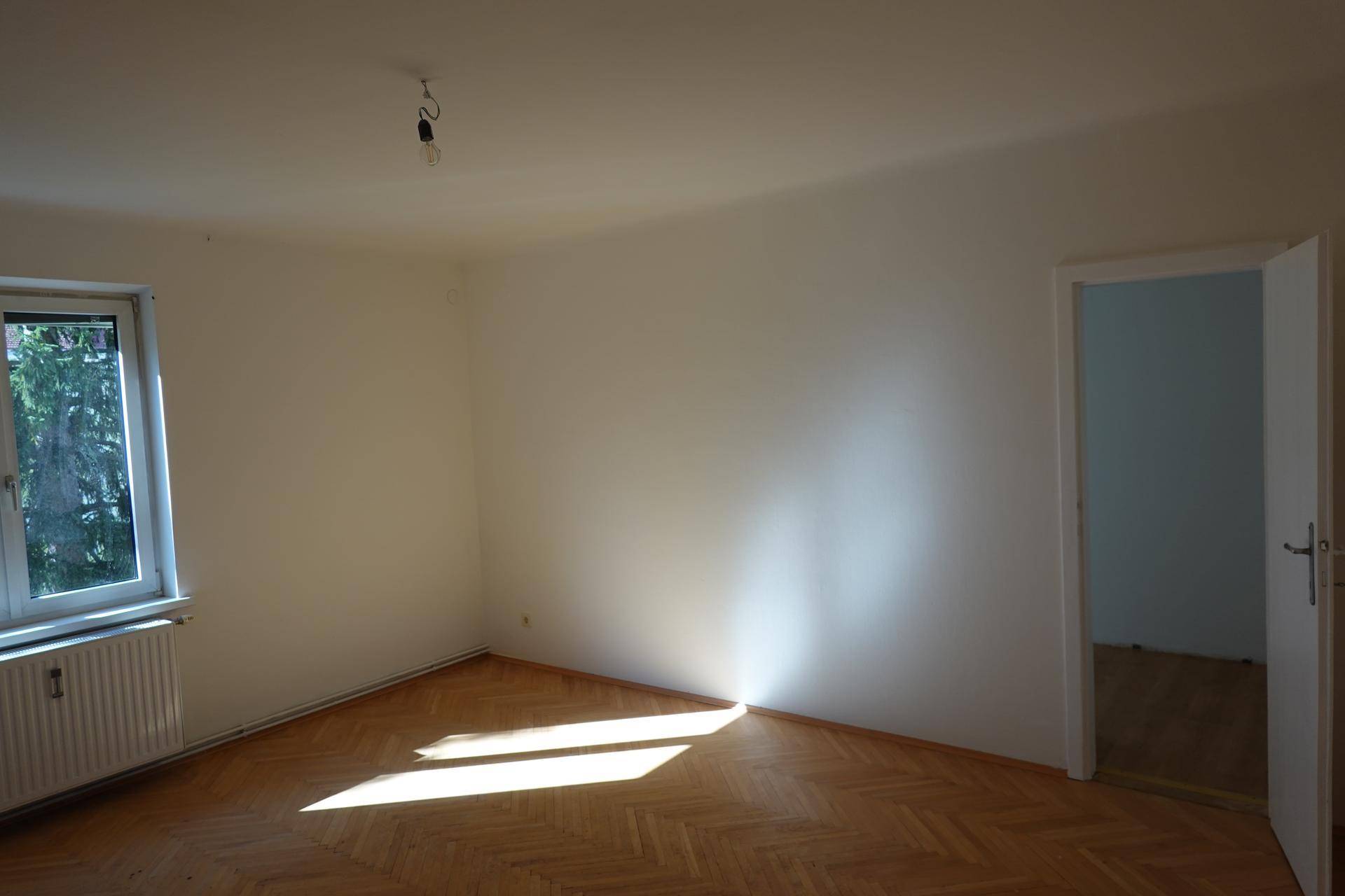 Heller Raum mit Fischgrätparkett, Fenster und Sonneneinfall, der den Boden beleuchtet.