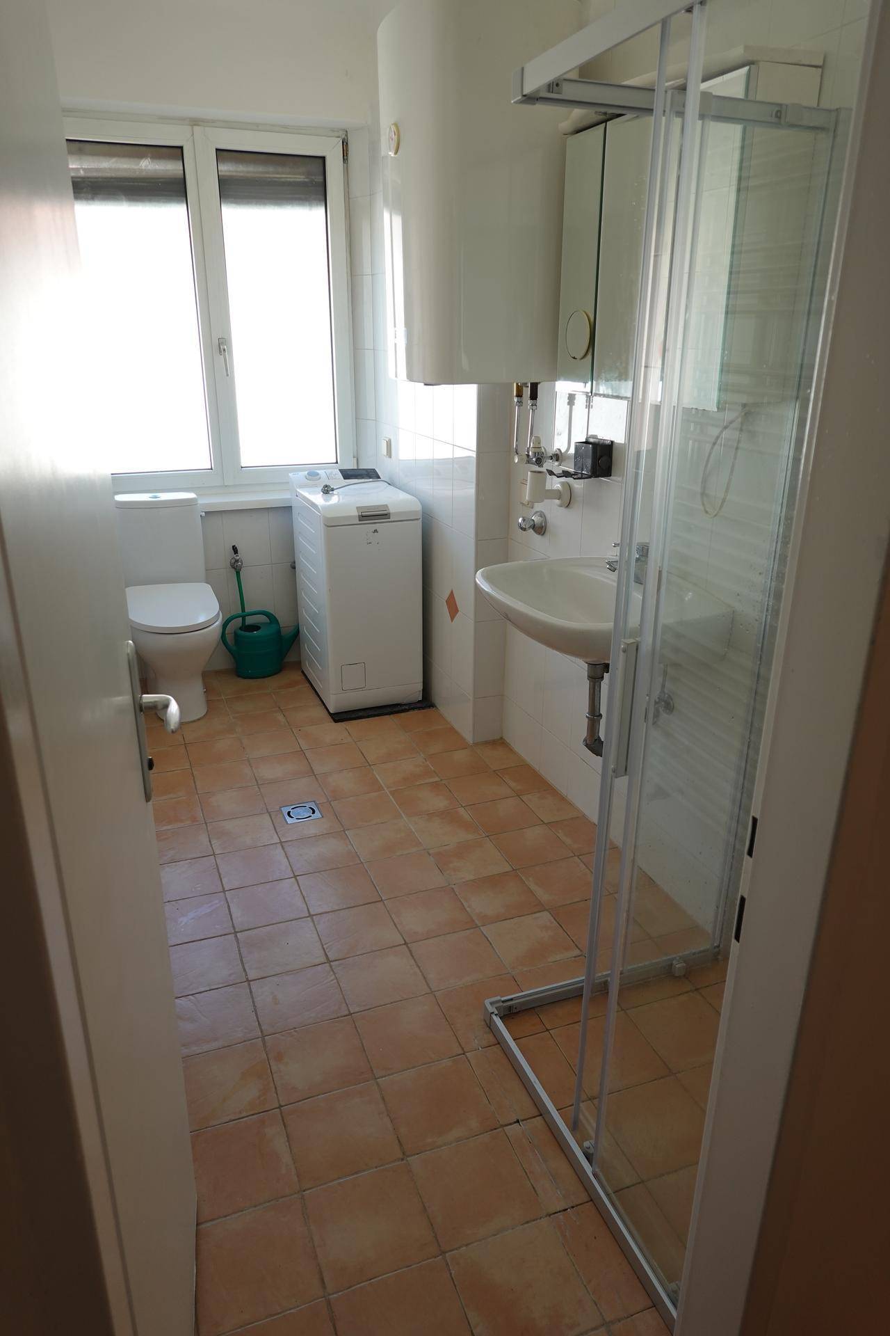 Badezimmer mit Fenster, WC, Waschmaschine, Waschbecken und Duschkabine mit Terrakottafliesen.