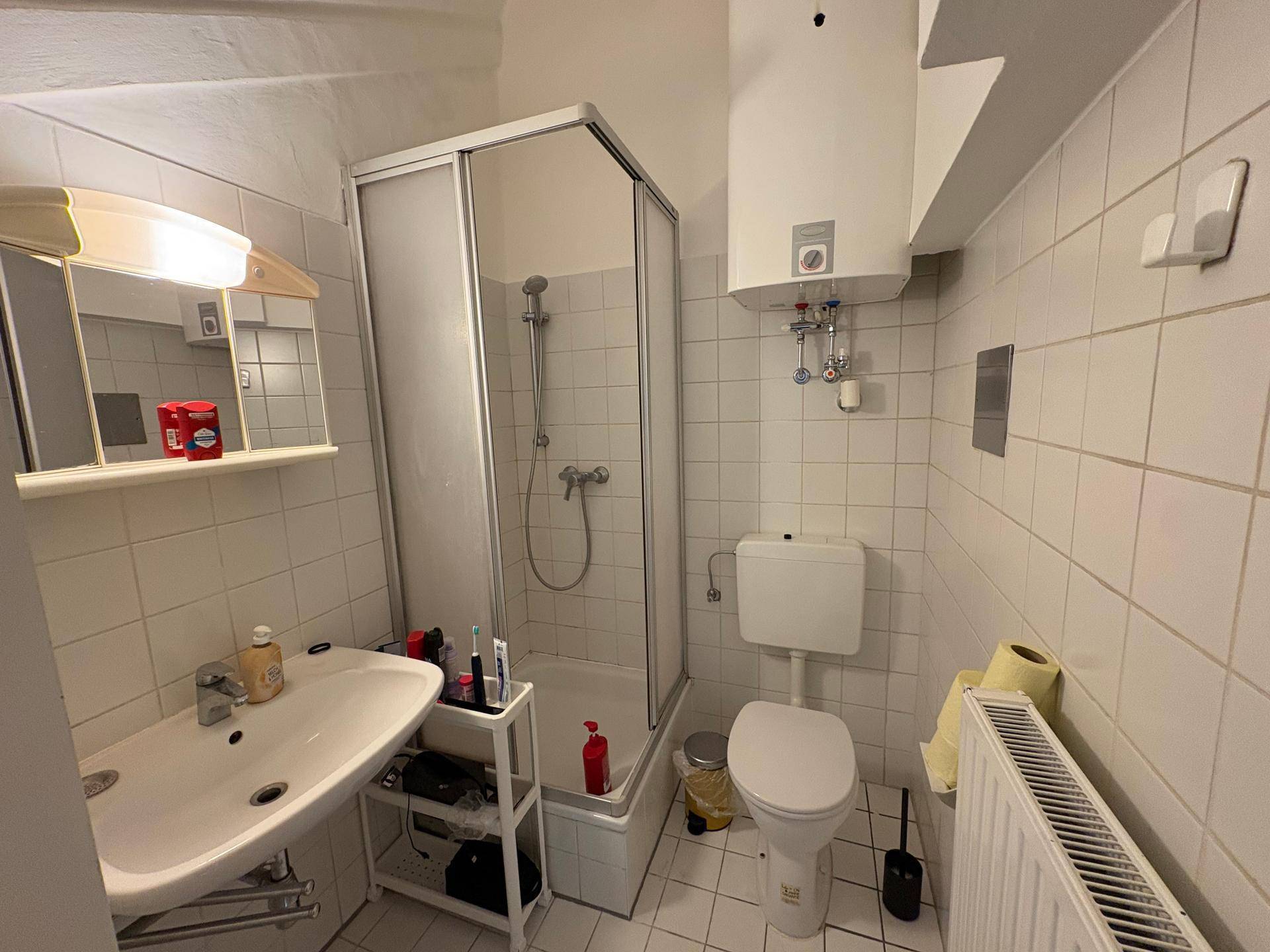 Einfaches Badezimmer mit Duschkabine, Toilette, Waschbecken und weißen Fliesen.