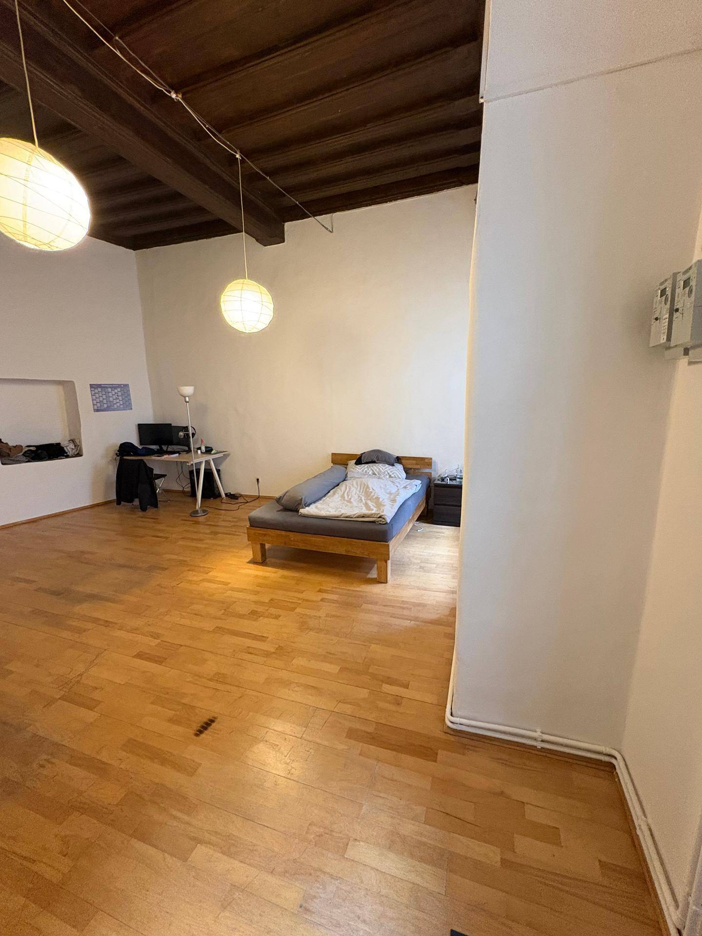 Heller Wohnraum mit Holzbalkendecke, Parkettboden und einem einfachen Bett in der Ecke.