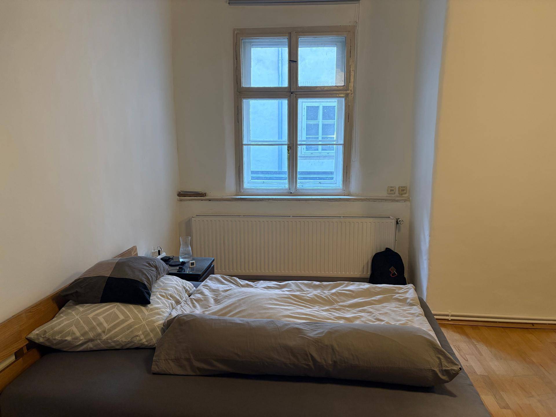 Schlafbereich mit einem einfachen Holzbett, Heizkörper unter dem Fenster und Parkettboden.