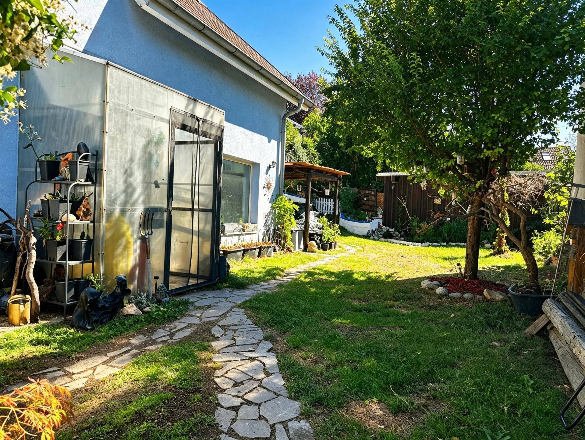 Grüner Gartenbereich mit Rasen, Bäumen, Steinpfad und einem Gewächshaus neben dem Haus.