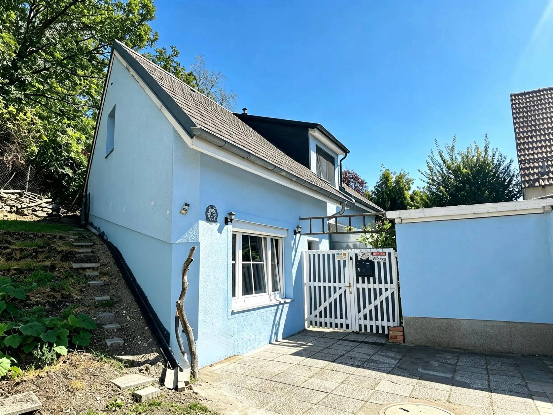 Hellblaues Haus mit weißem Gartentor und gepflastertem Zugang unter blauem Himmel.