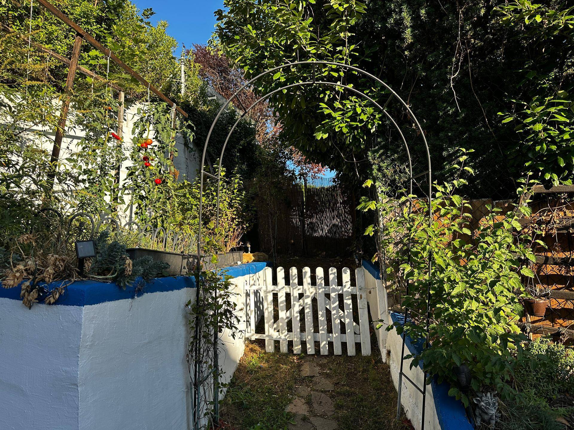 Gartenweg führt durch einen Bogen, flankiert von Hochbeeten und einem weiteren weißen Gartentor.