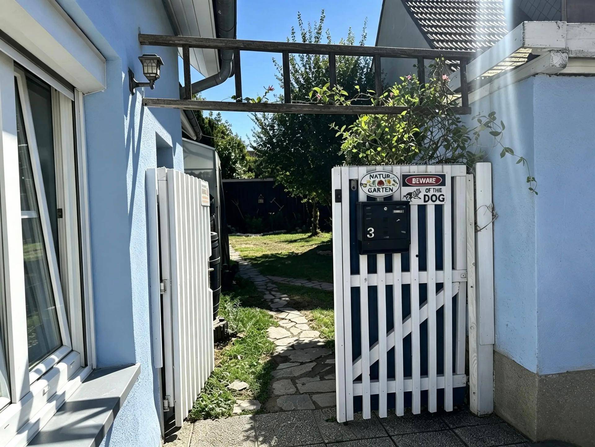 Eingangsbereich mit weißem Gartentor, Briefkasten und einem Pfad, der in den Garten führt.