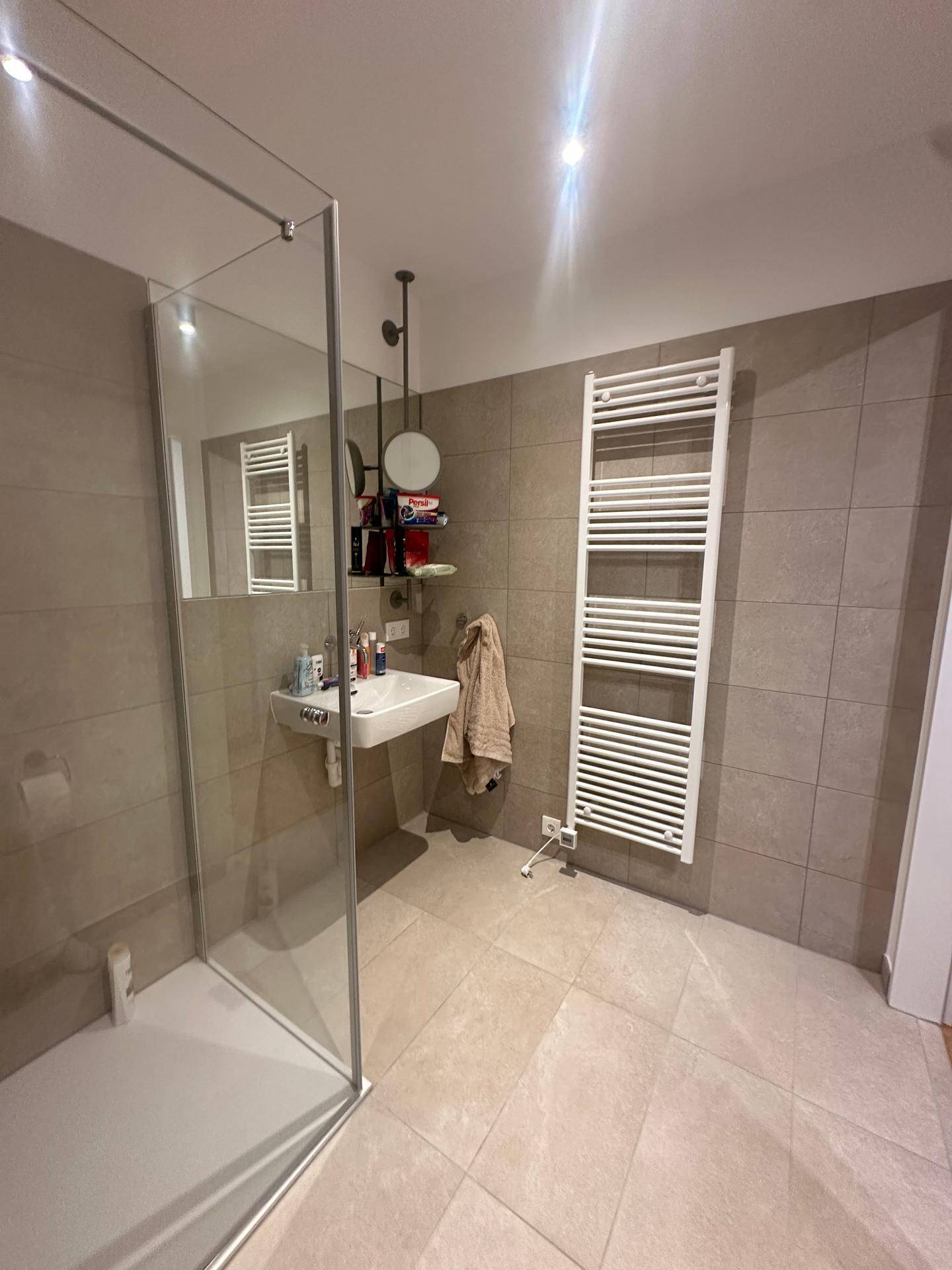 Funktionales Badezimmer mit Duschkabine, Waschbecken und beheiztem Handtuchhalter auf hellen Fliesen.