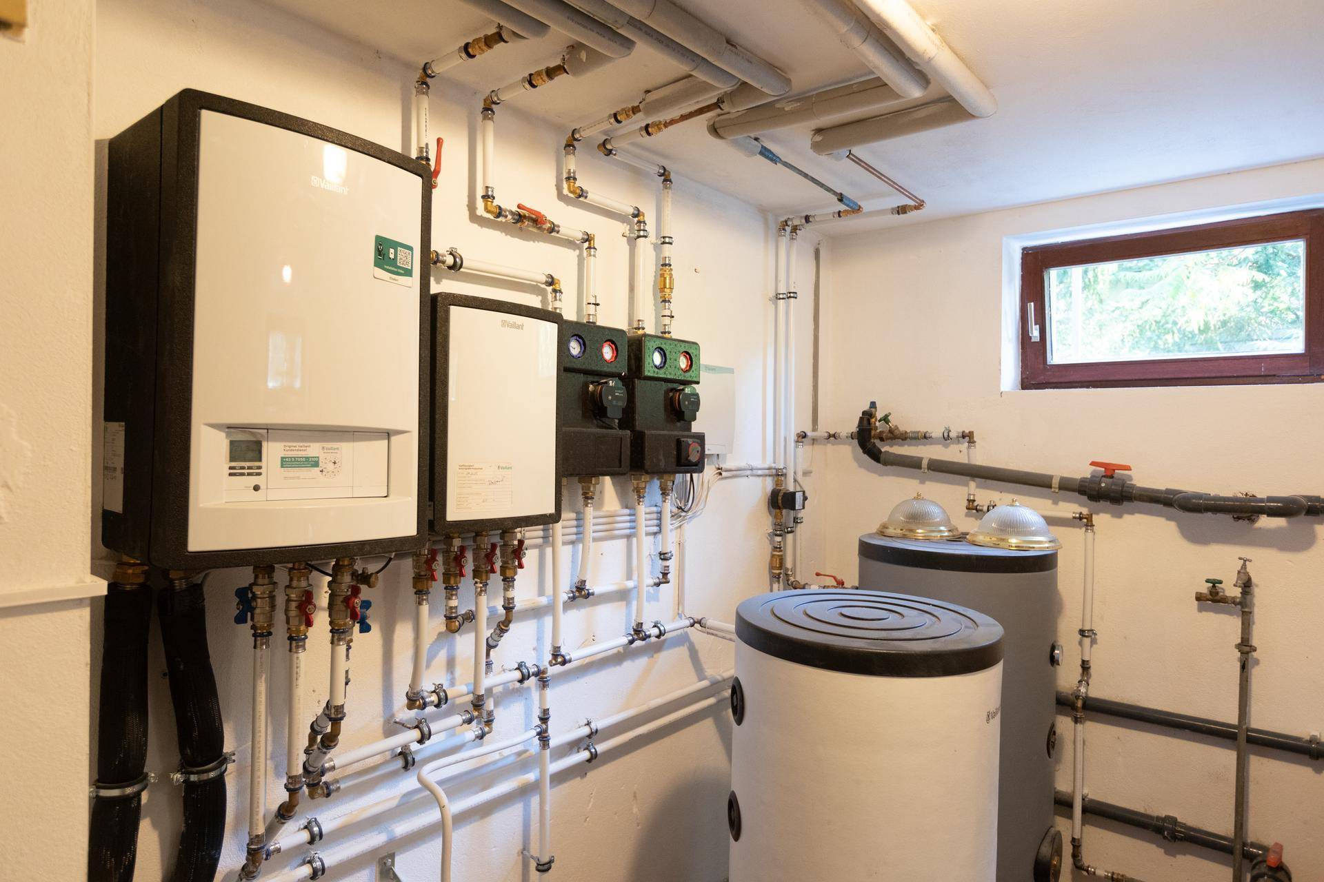 Technikraum im Keller mit Heizungsanlage, Warmwasserspeichern und sichtbaren Rohrleitungen.