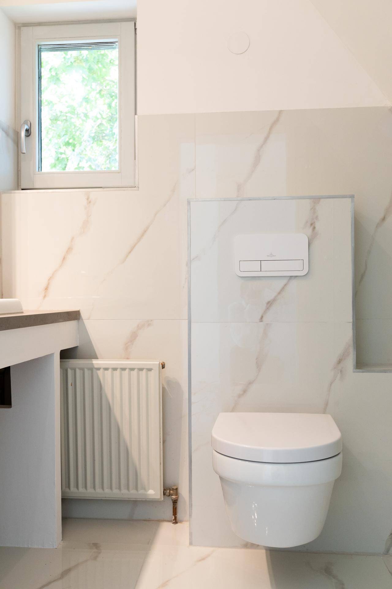 Modernes Badezimmer mit wandhängendem WC, Fenster, Heizkörper und Marmor-Effekt-Fliesen.