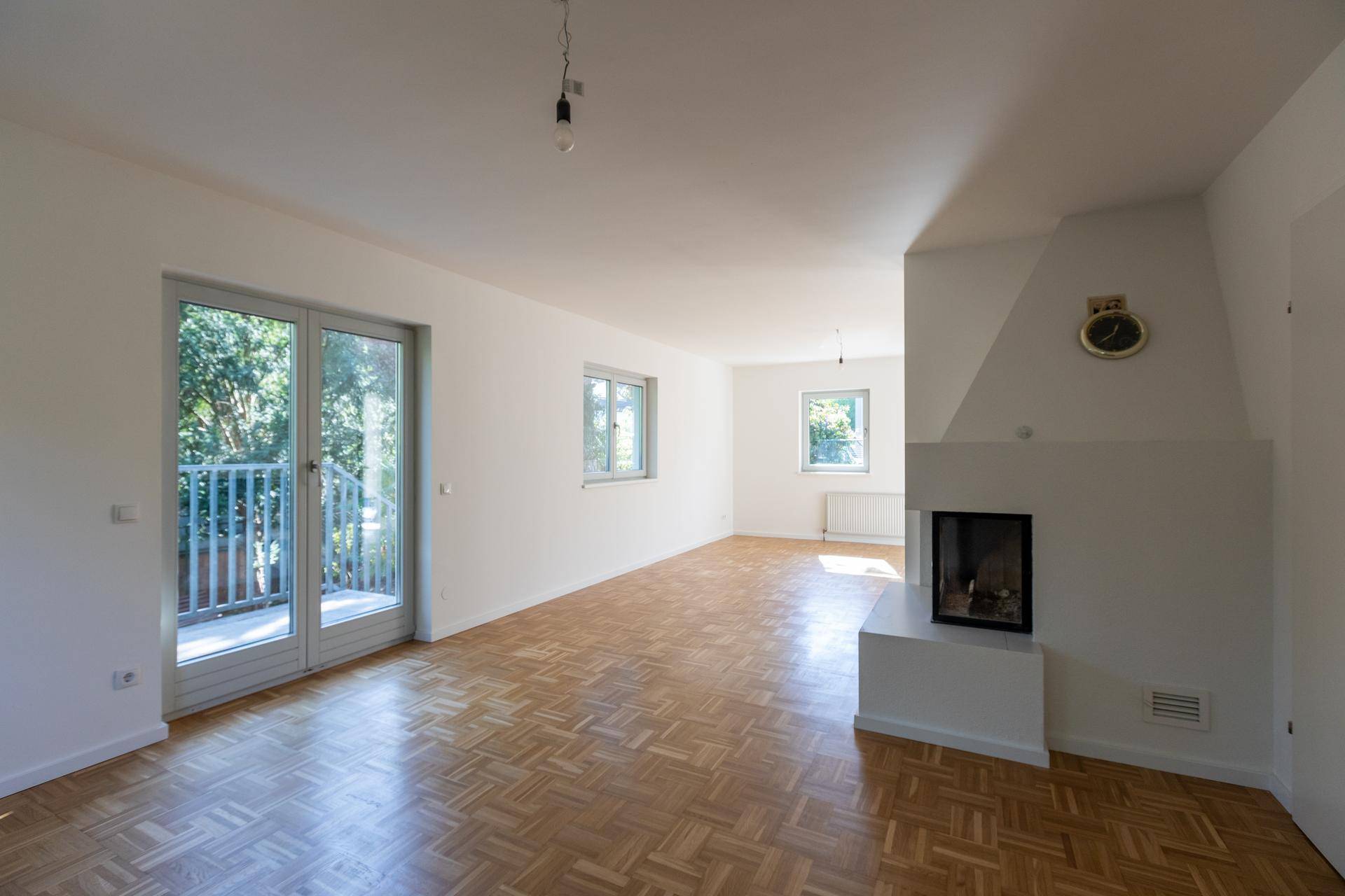 Helles Wohnzimmer mit Fischgrätparkett, Kamin und Zugang zu einem Balkon mit Blick ins Grüne.