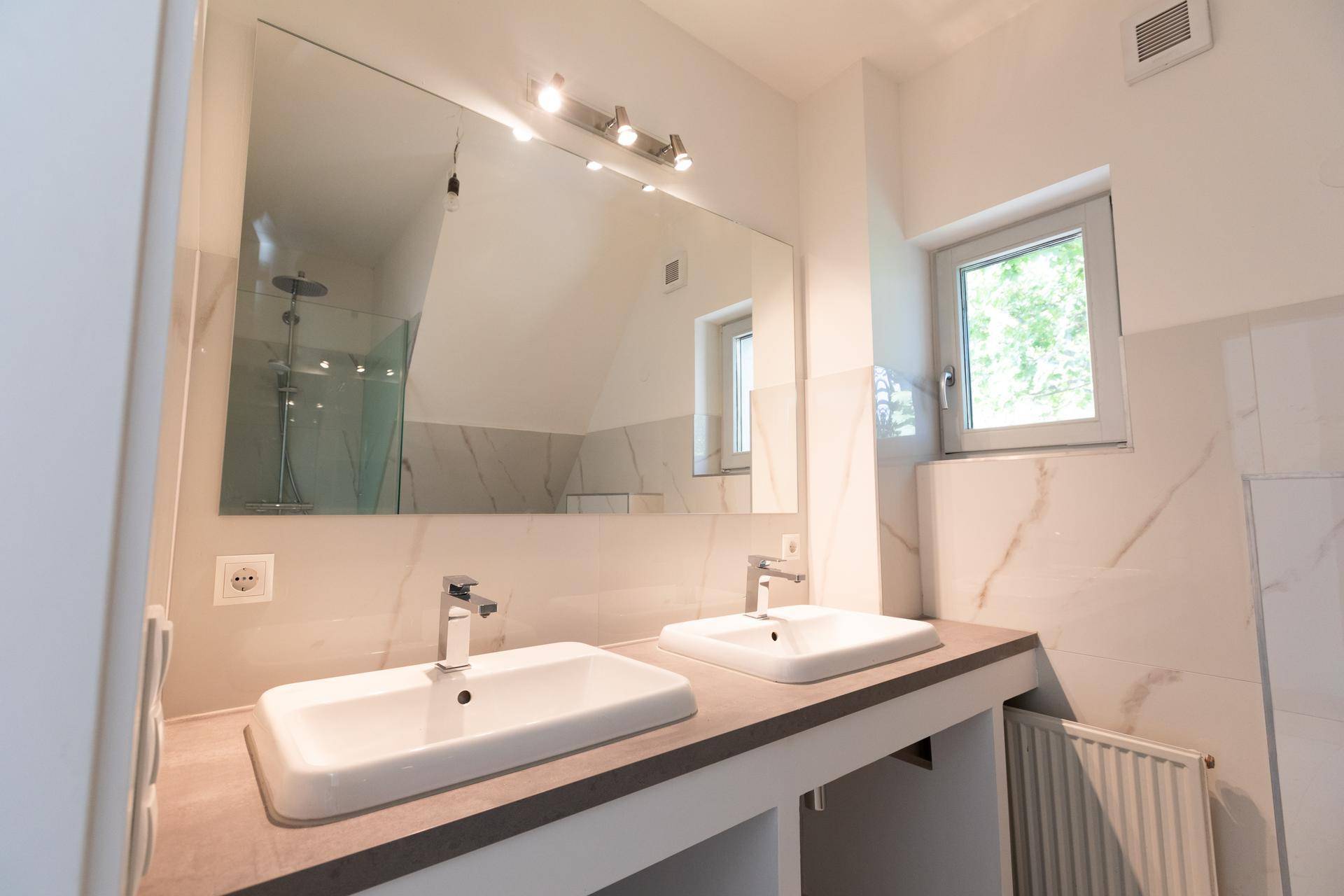 Helles Badezimmer mit Doppelwaschbecken, großem Spiegel, Fenster und modernen Marmorfliesen.