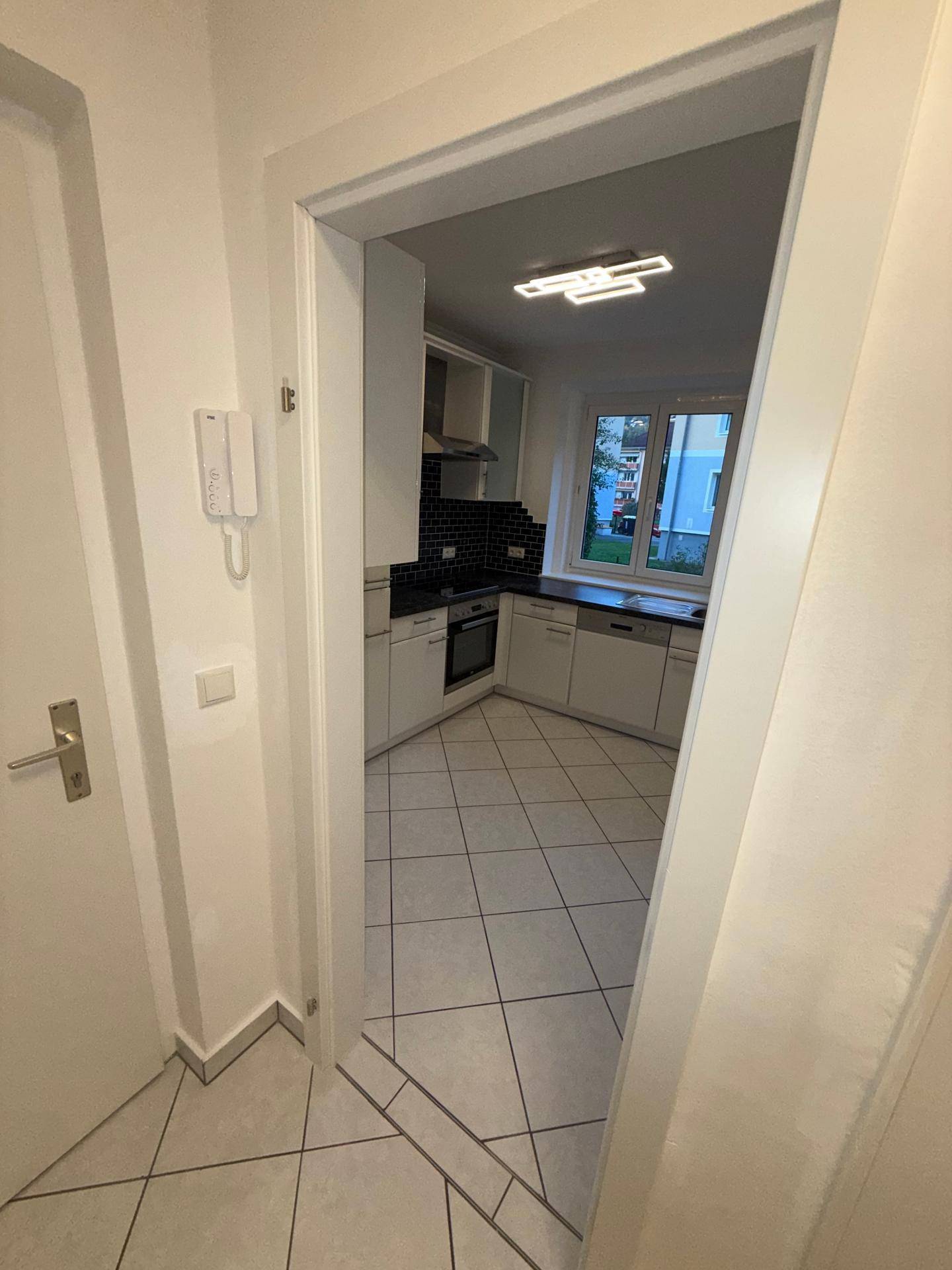 Blick vom Flur in die Küche mit weißen Schränken, schwarzem Fliesenspiegel und Fenster.
