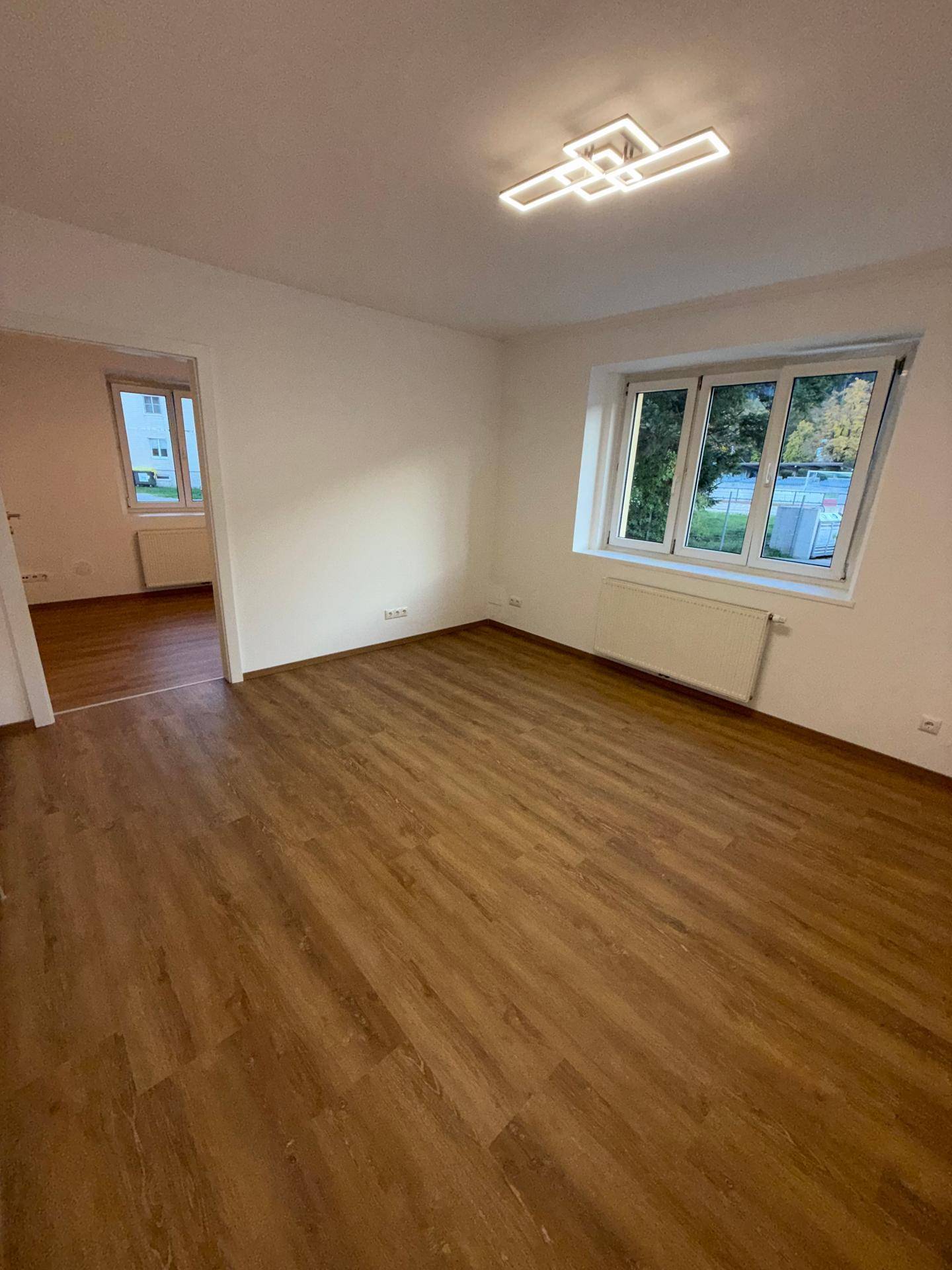 Freundliches Zimmer mit Laminatboden, weißen Wänden und Blick in einen weiteren Raum.