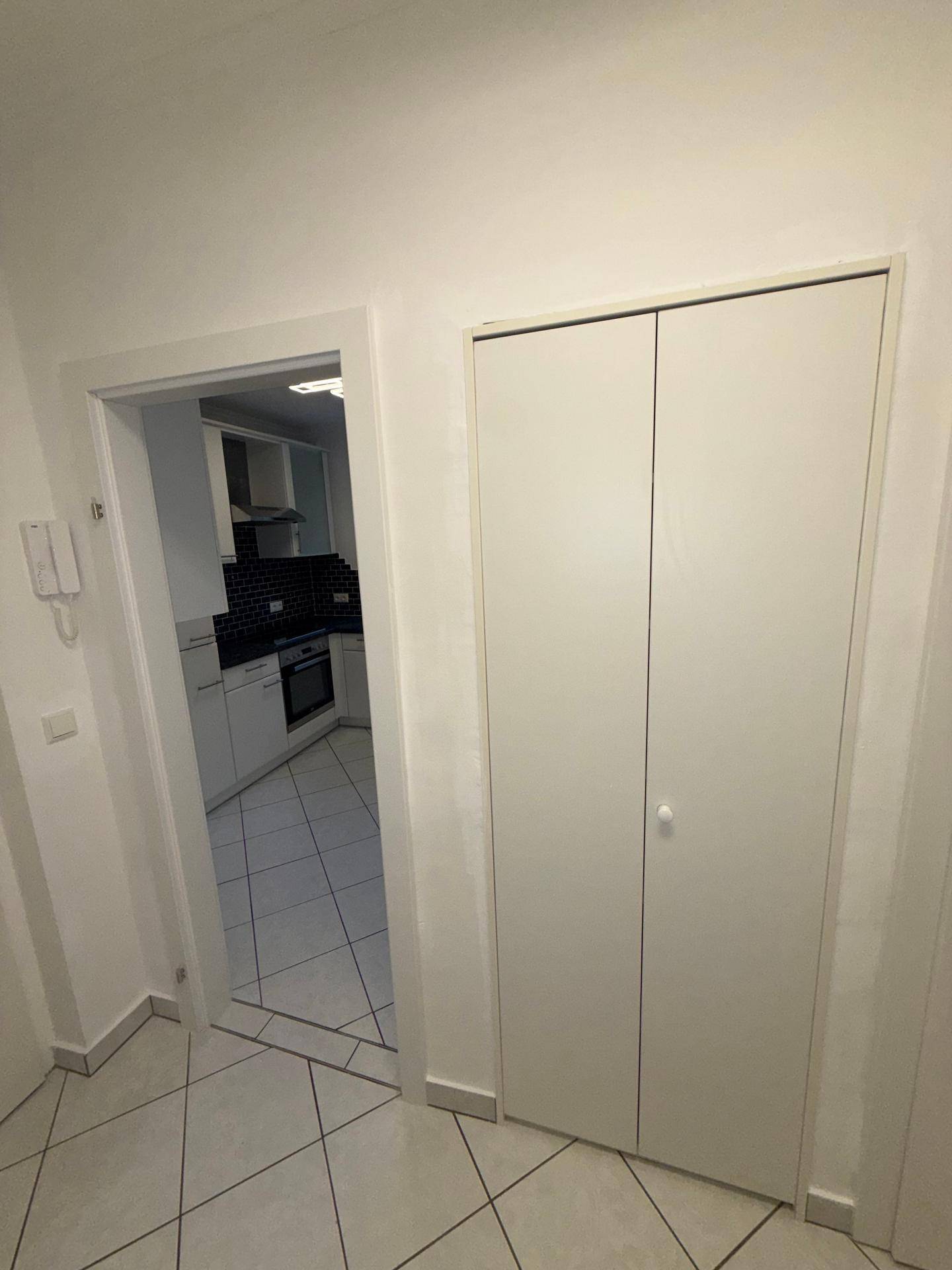 Heller Flur mit Fliesenboden und Zugang zu einer modernen Küche mit dunklem Fliesenspiegel.