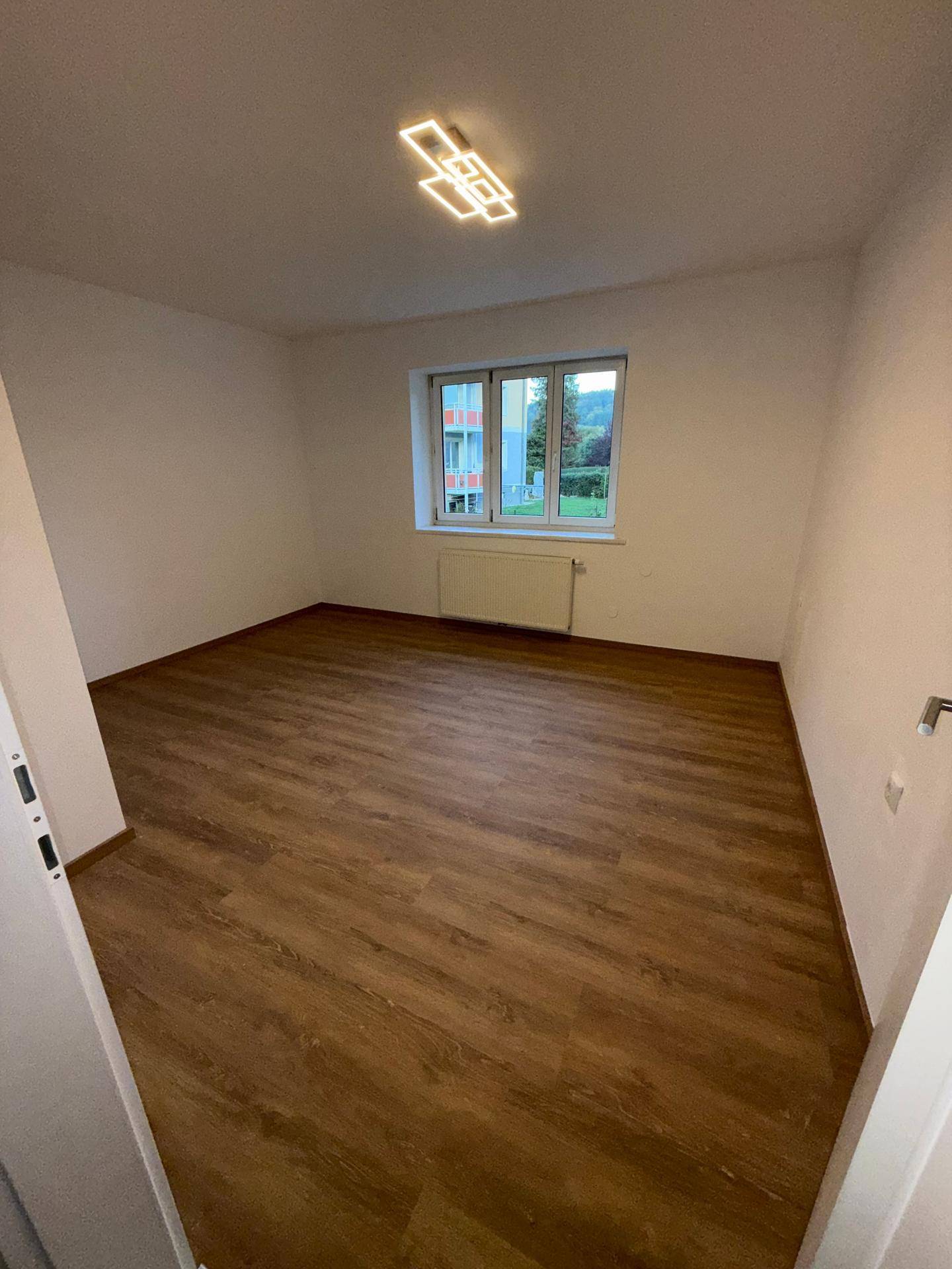 Geräumiges Zimmer mit Laminatboden, weißen Wänden und einem Fenster mit Blick auf die Umgebung.