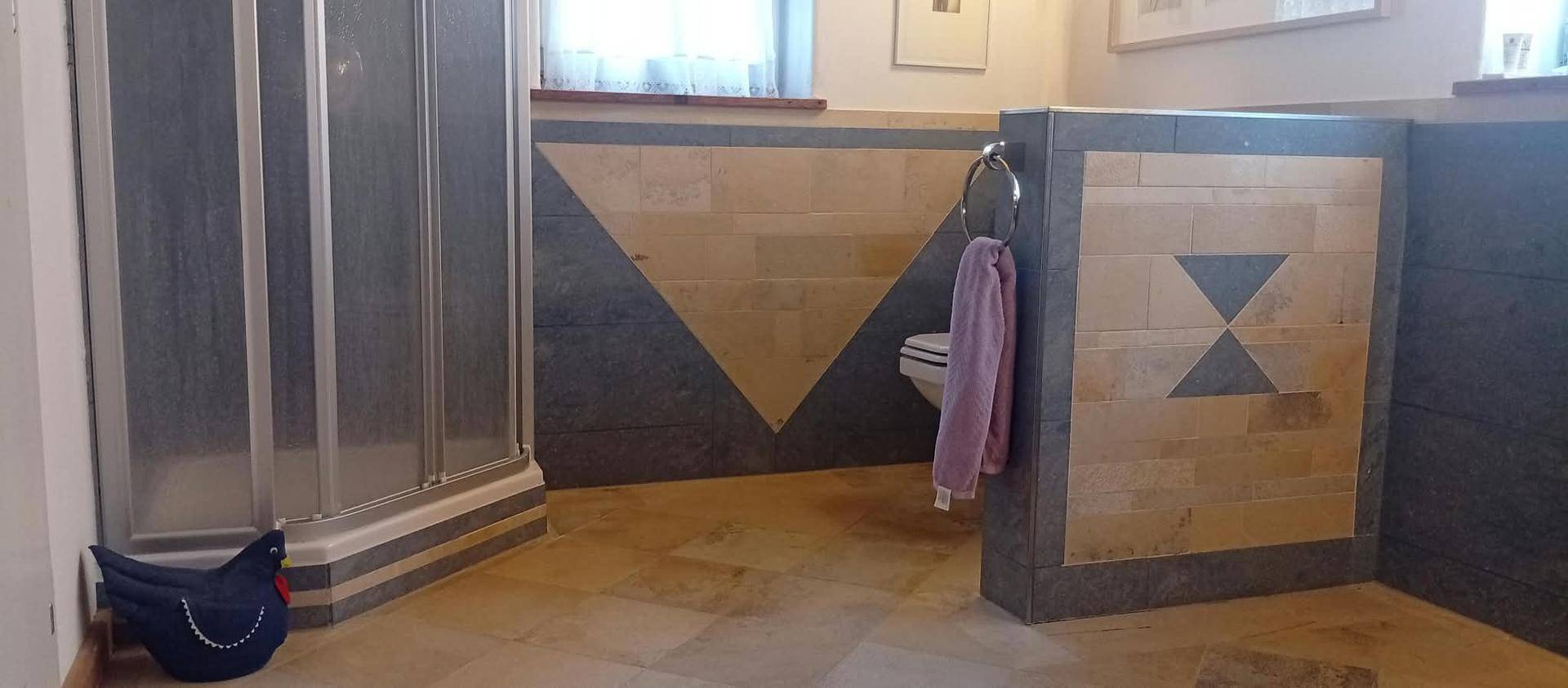 Badezimmer mit Duschkabine, WC hinter einer Trennwand und einem Fenster für natürliches Licht.