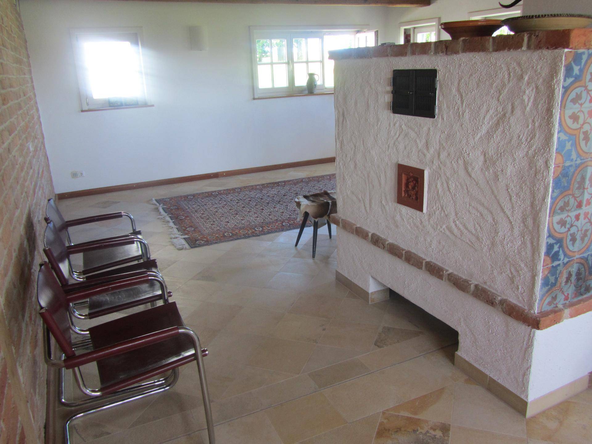 Heller Raum mit Fliesenboden, Kachelofen und Sitzgelegenheiten, offen zum Wohnbereich.