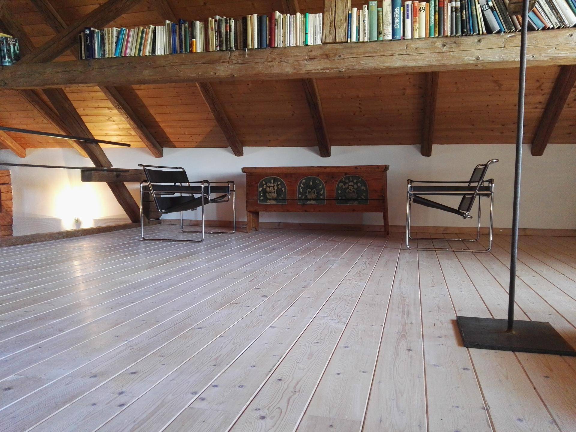 Helles Dachzimmer mit schrägen Holzdecken, Holzdielenboden und einer Bücherablage auf dem Balken.