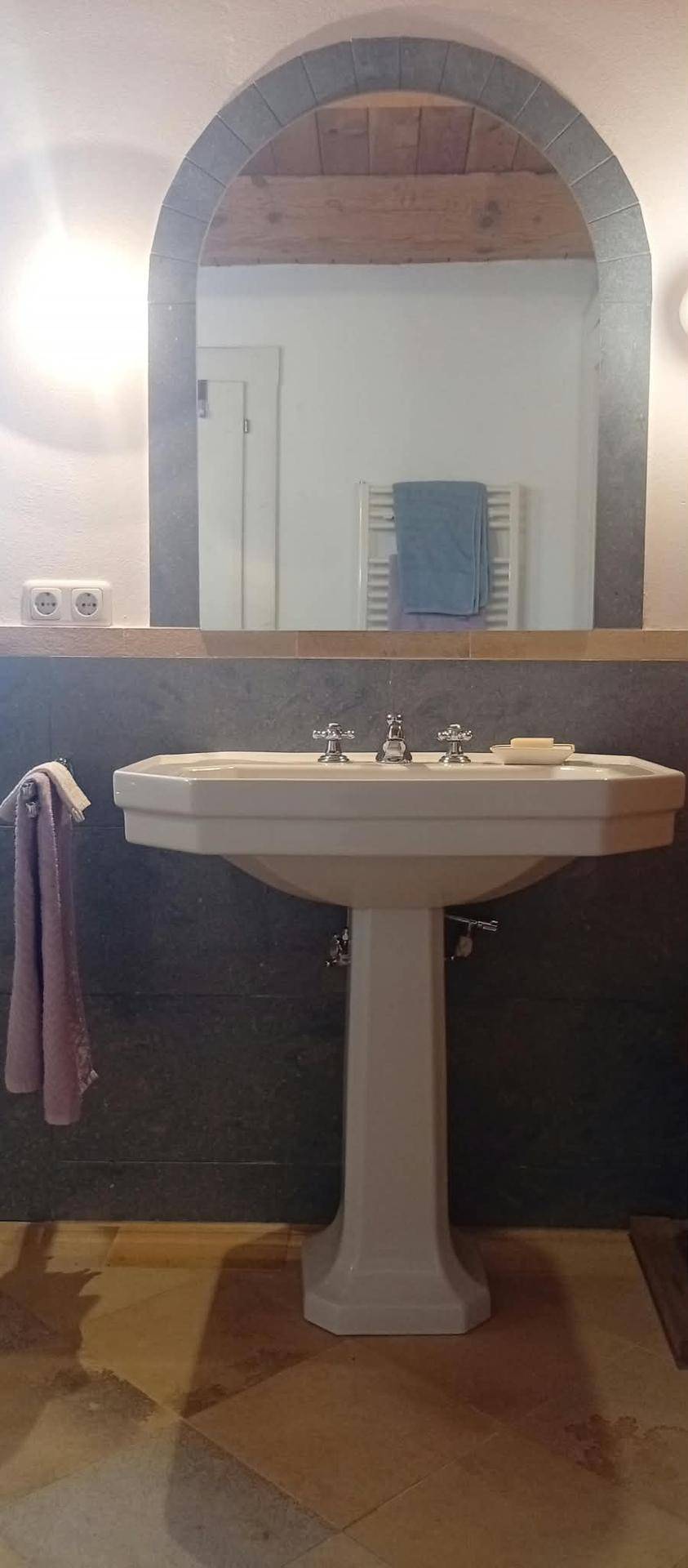 Badezimmer mit Standwaschbecken, gewölbtem Spiegel und Holzbalkendecke, inklusive Handtuchheizkörper.