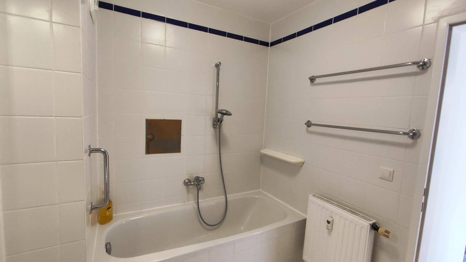 Badewanne mit Duschfunktion in einem gefliesten Badezimmer, ergänzt durch Handtuchhalter und Heizkörper.