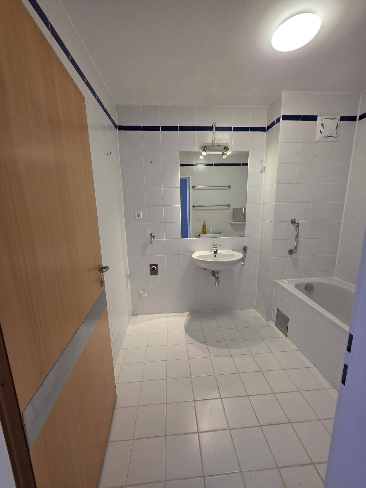 Badezimmer mit weißen Fliesen, Badewanne, Waschbecken und Spiegel, ausgestattet mit Haltegriffen.