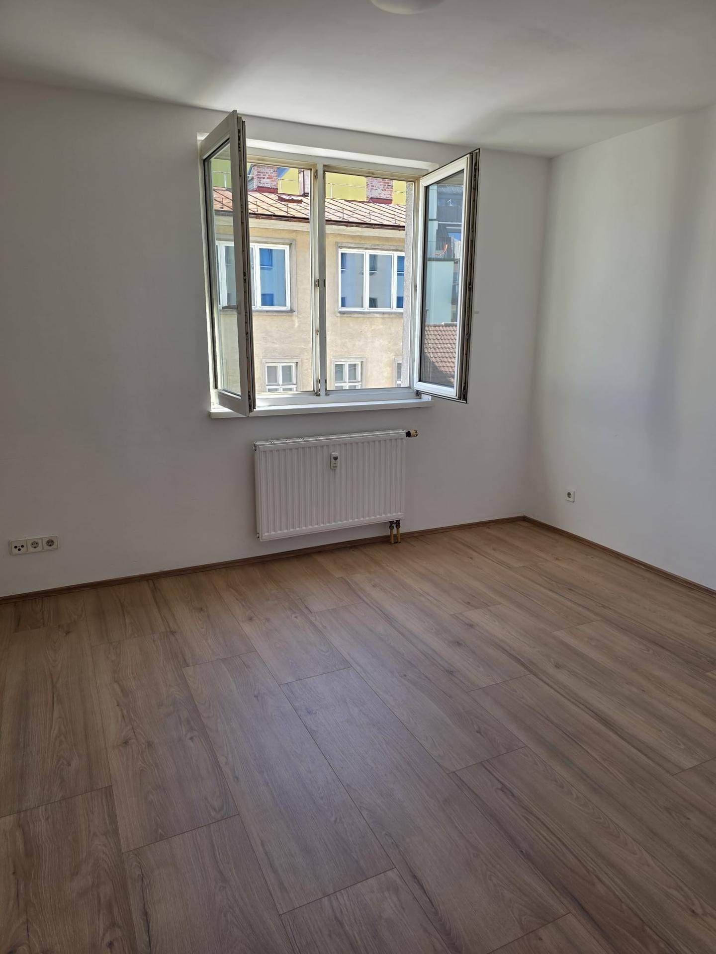 Geräumiges Schlafzimmer mit Laminatboden und einem Fenster, das einen Blick auf die Nachbargebäude bietet.