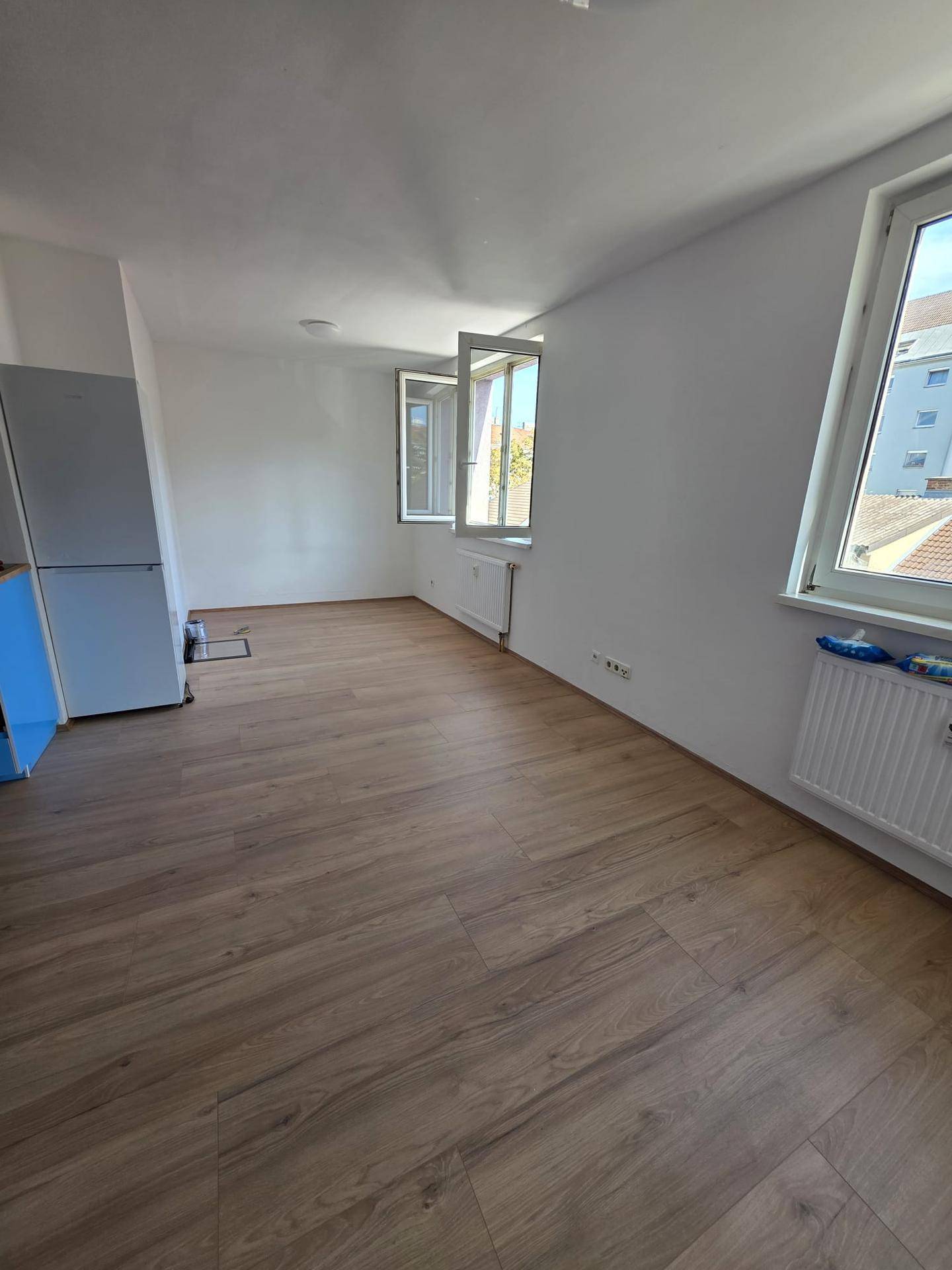 Heller Wohnbereich mit Laminatboden und mehreren Fenstern, die viel Tageslicht hereinlassen.