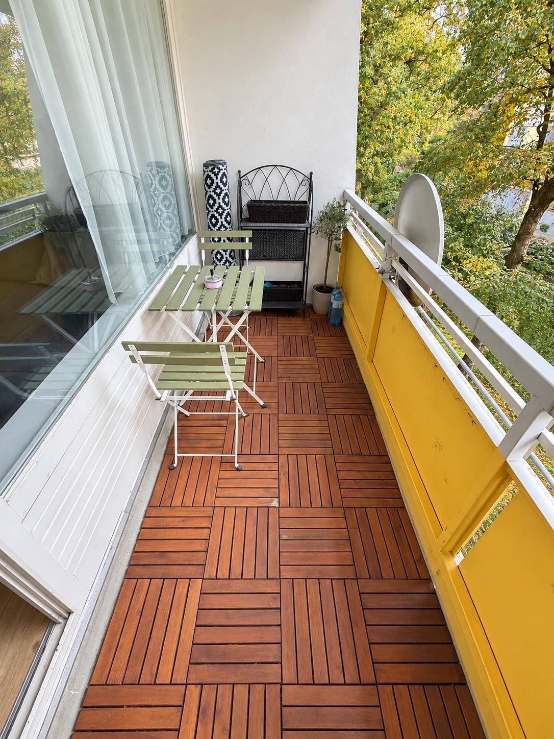 Kleiner Balkon mit Holzbodenfliesen und Sitzgelegenheiten, umgeben von grünen Bäumen.