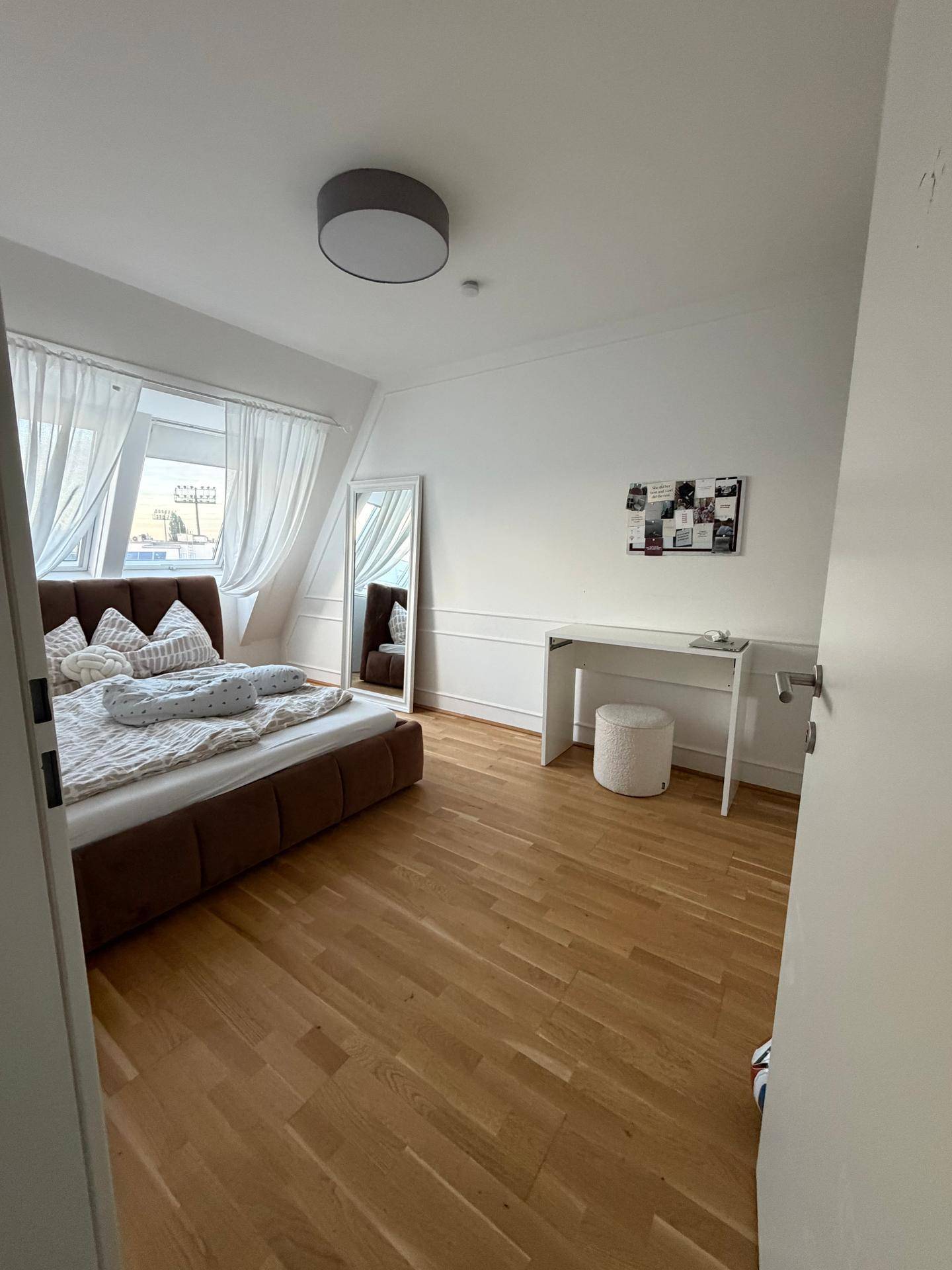 Helles Schlafzimmer mit Holzboden, großem Spiegel und einem Arbeitsbereich am Fenster.