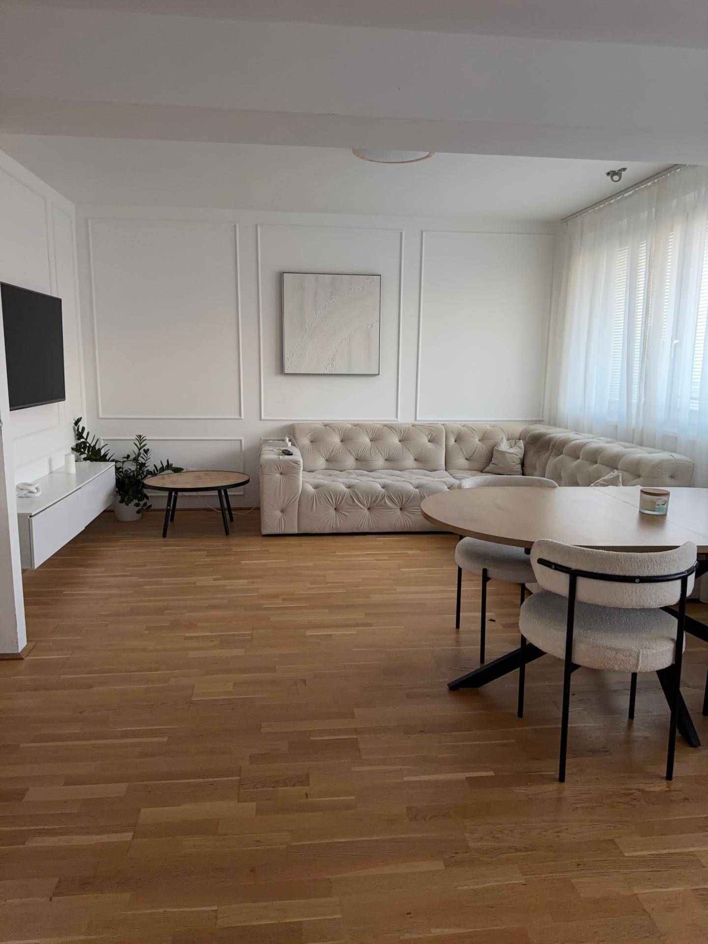 Heller Wohnbereich mit beigem Sofa, Fernseher und einem großen Esstisch im Vordergrund.