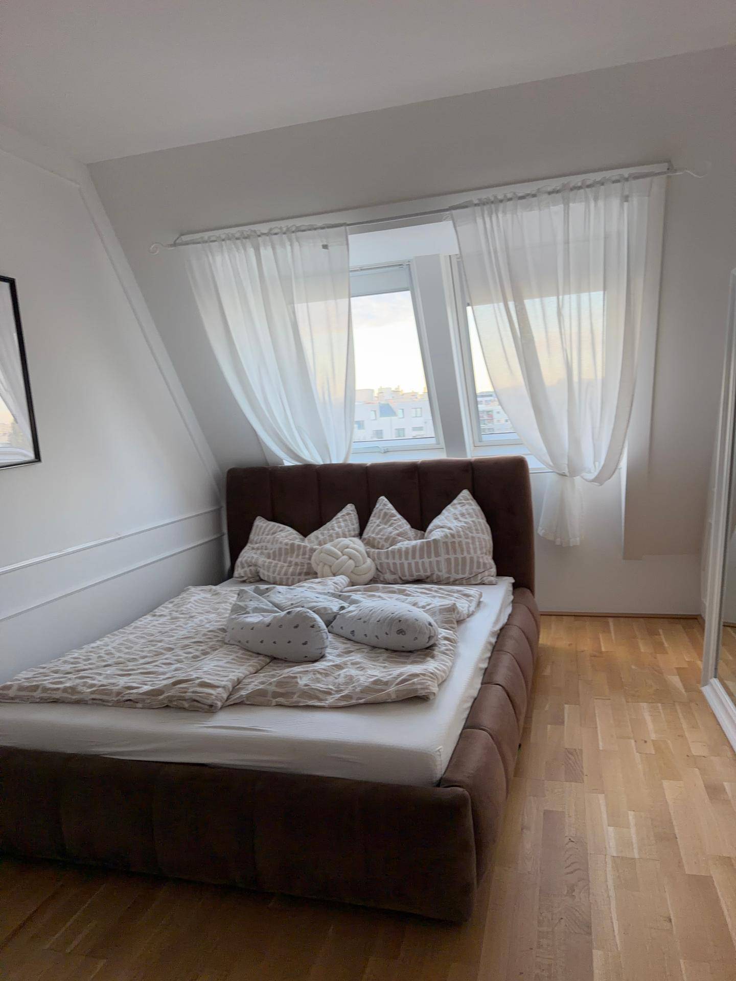 Gemütliches Schlafzimmer mit braunem Polsterbett, Holzboden und Dachschräge am Fenster.