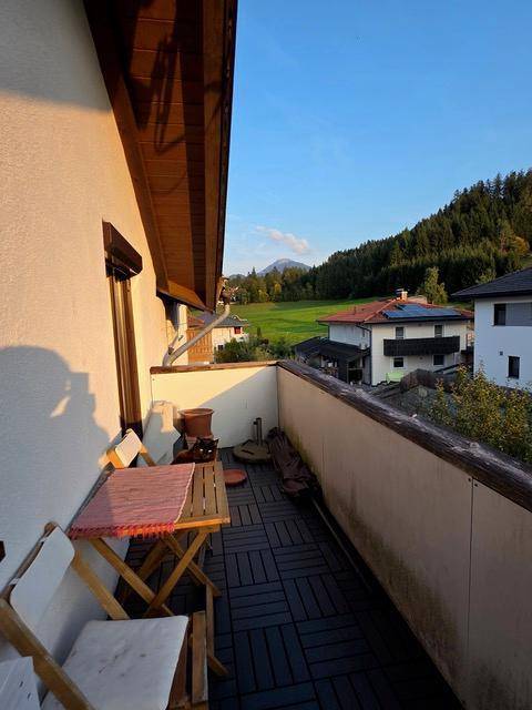 Sonniger Balkon mit Holztisch und Stühlen, umgeben von grüner Landschaft und Bergen.