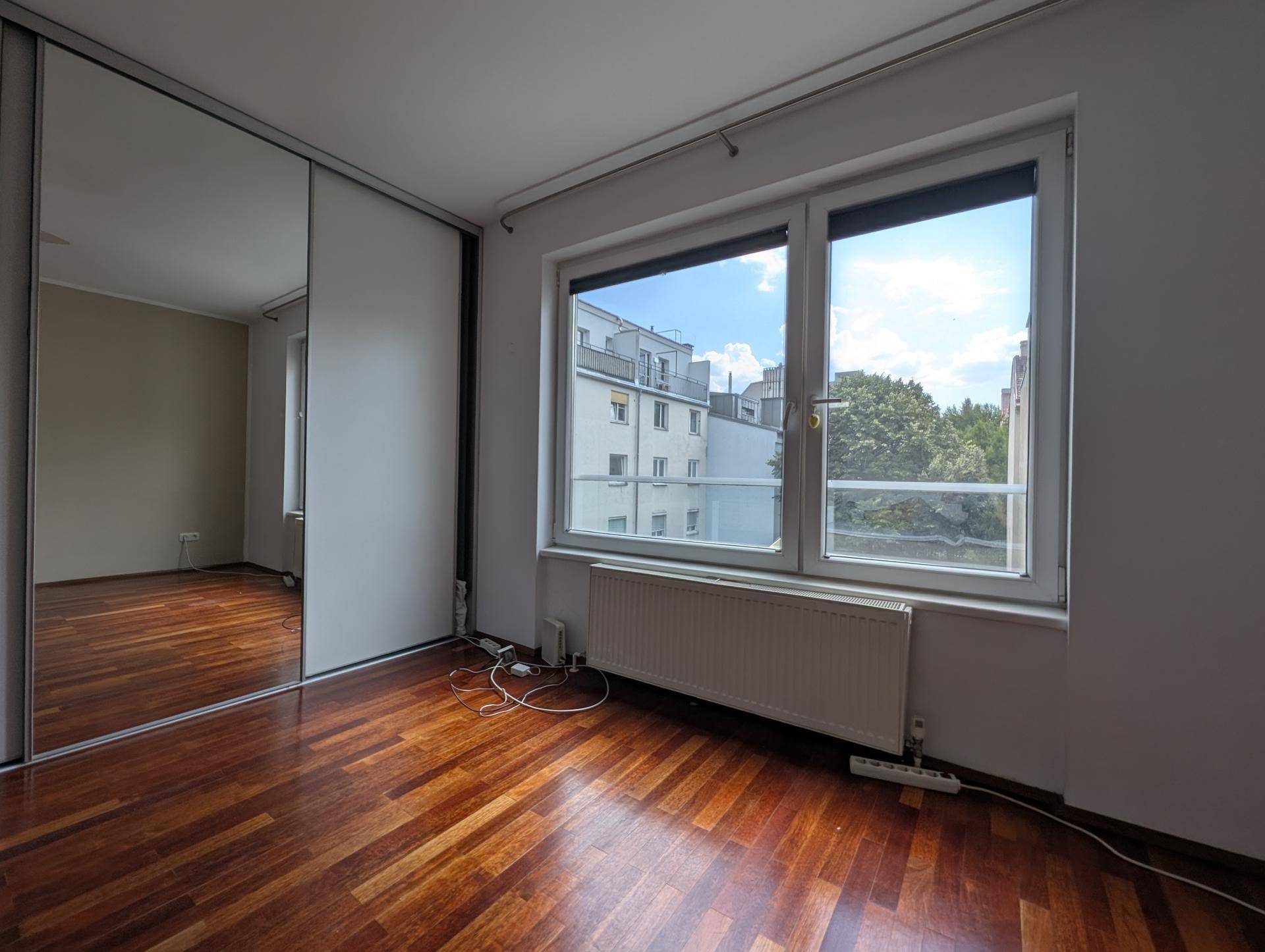 Schlafzimmer mit Parkettboden, großem Fenster und einem Spiegelschrank an der Wand, der den Raum erweitert.