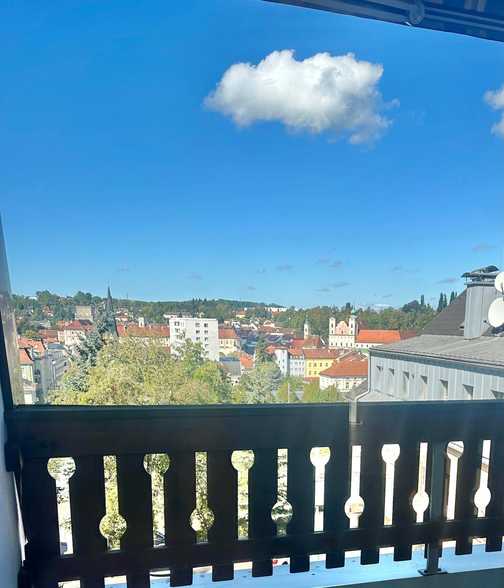 Weitblick von einem Balkon über eine Stadtlandschaft mit blauem Himmel und einer Wolke.