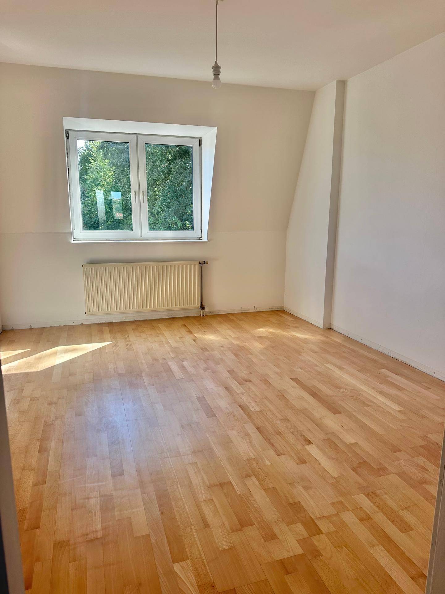 Helles Schlafzimmer mit Holzfußboden und einem Fenster, das einen Blick ins Grüne bietet.