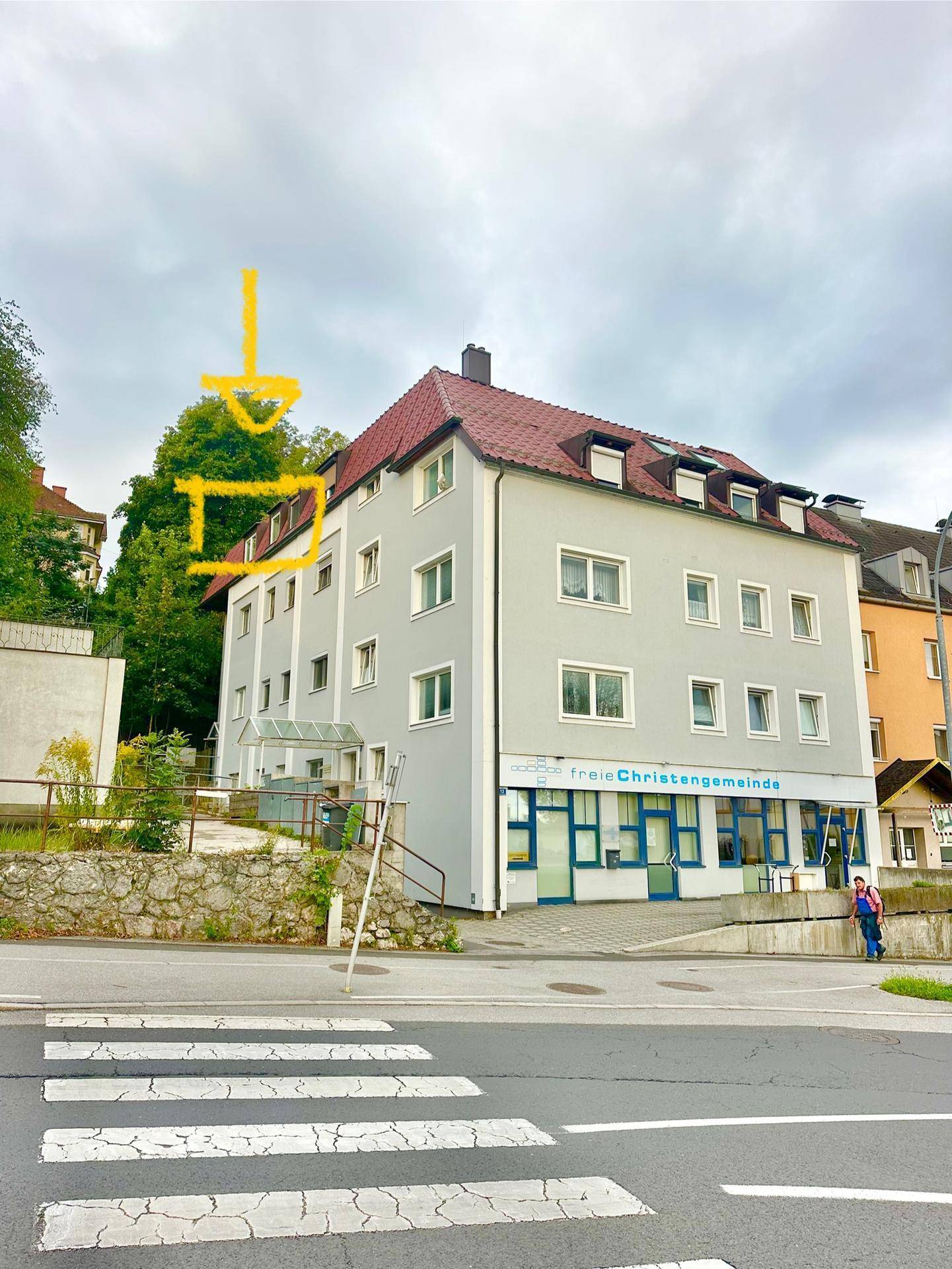 Mehrstöckiges Wohngebäude mit Satteldach und grauer Fassade an einer Straße, mit Markierungen.