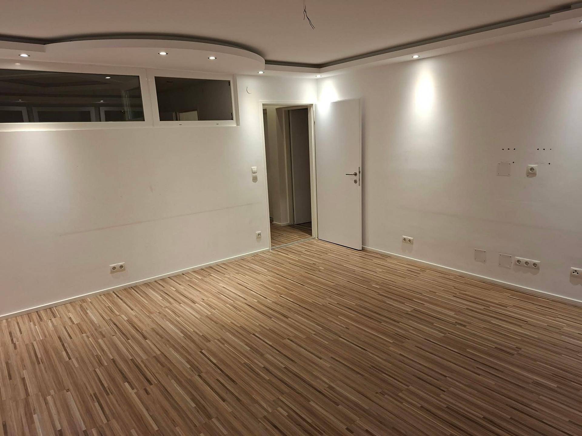 Geräumiges Wohnzimmer mit Laminatboden, moderner Deckenbeleuchtung und Zugang zu einem weiteren Raum.