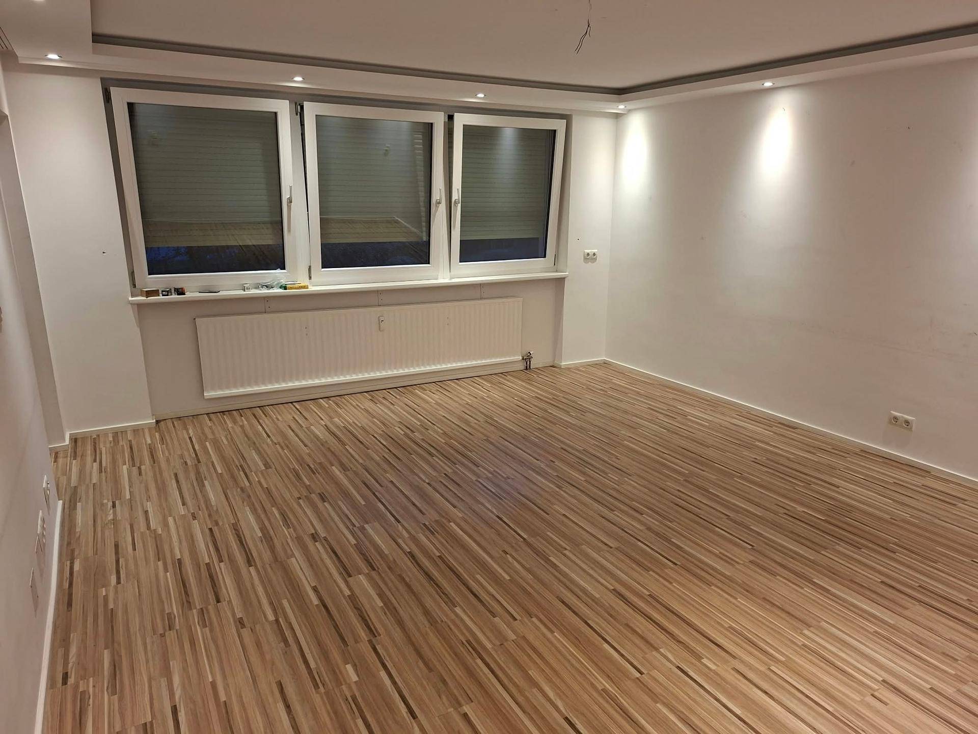 Geräumiges Wohnzimmer mit Laminatboden, großen Fenstern und integrierter Deckenbeleuchtung.