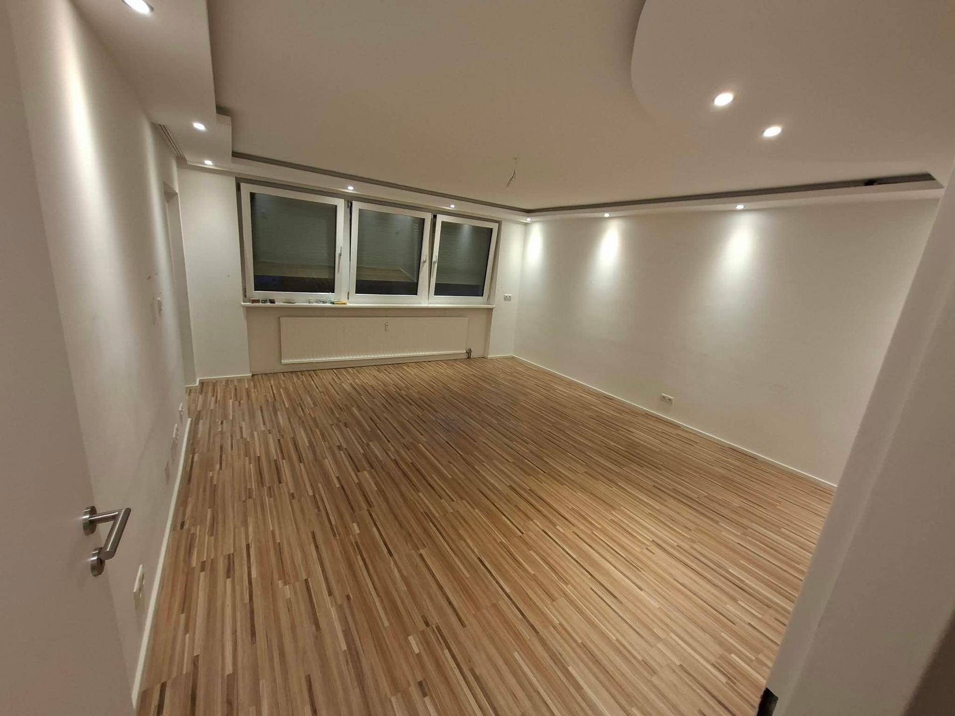 Geräumiges Wohnzimmer mit Laminatboden, großen Fenstern und moderner Deckenbeleuchtung.