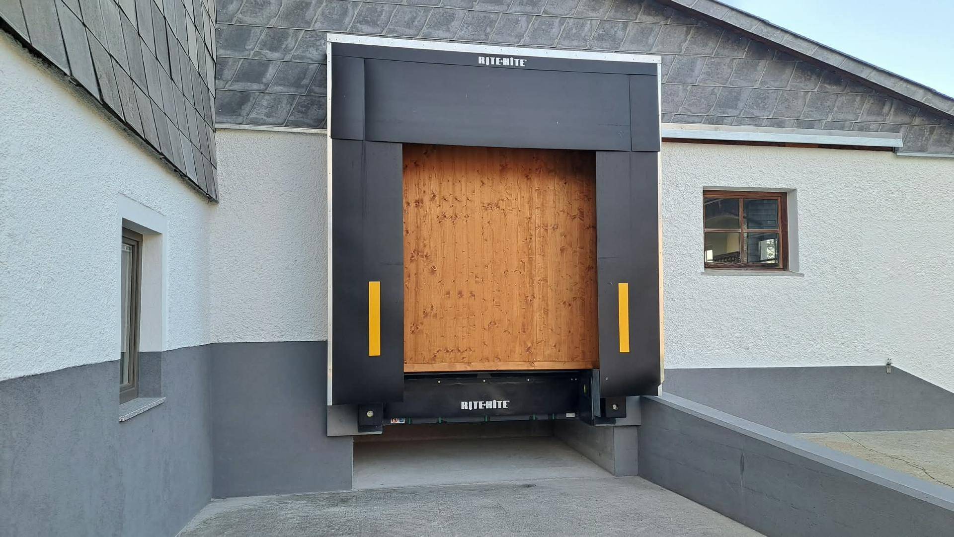 Außenansicht einer Laderampe mit schwarzer Mechanik und Holztor an einem Gebäude mit weiß-grauer Fassade.