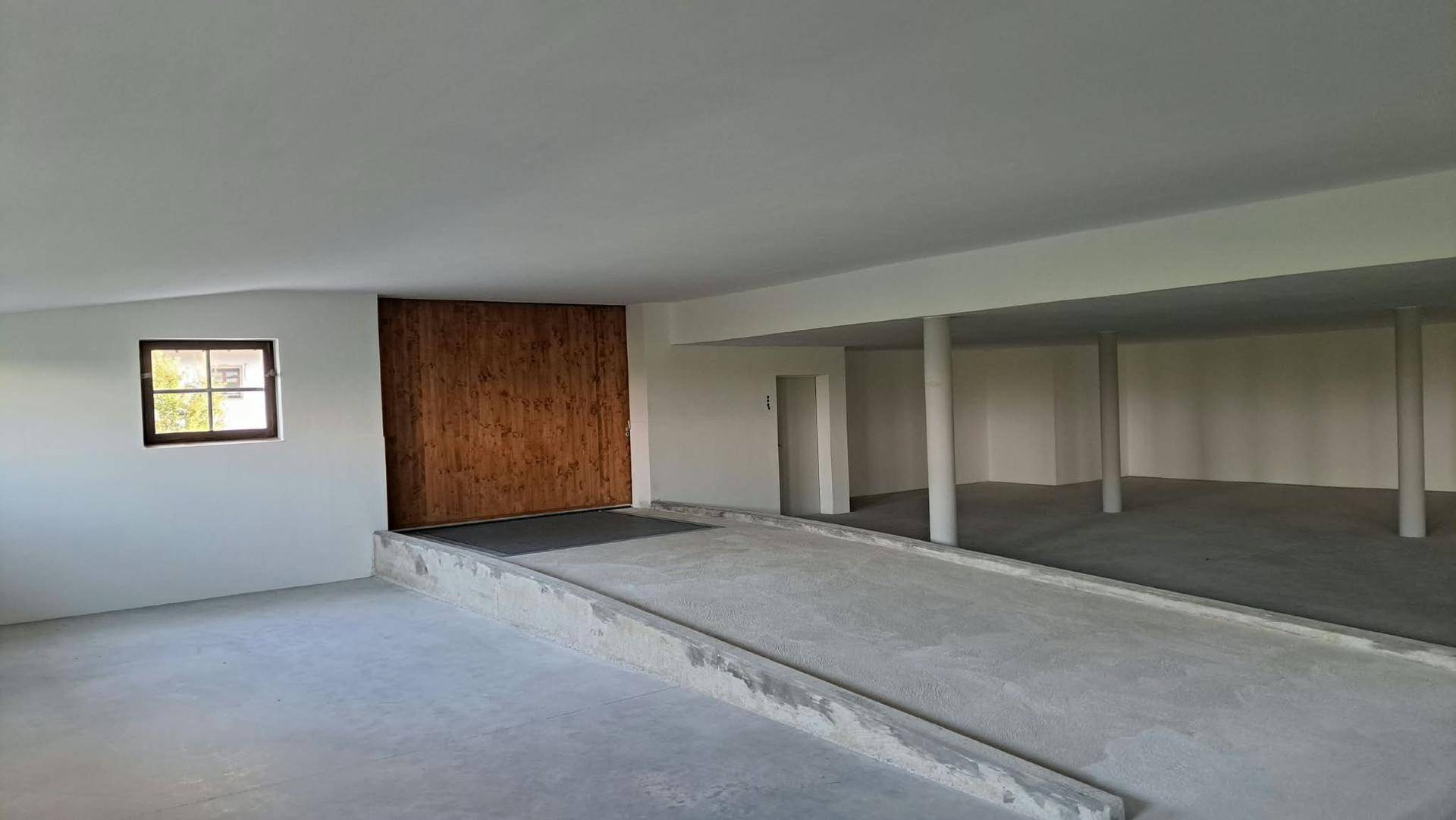 Großer leerer Innenraum mit Betonboden, weißen Wänden, einer Betonrampe und mehreren weißen Säulen.