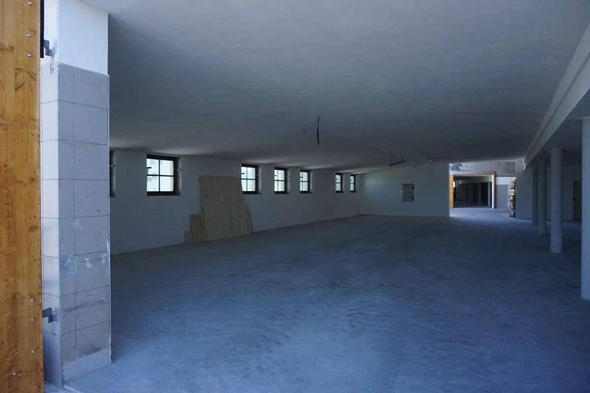 Großer, leerer Innenraum mit Betonboden, weißen Wänden und mehreren Säulen, eine Fensterreihe ist sichtbar.