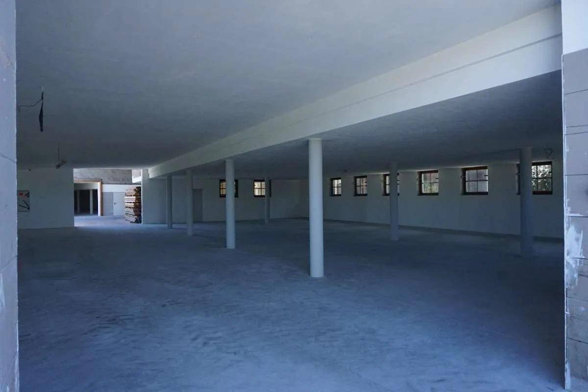 Großer, leerer Innenraum mit Betonboden, weißen Wänden und mehreren Säulen, kleine Fenster in der Ferne.