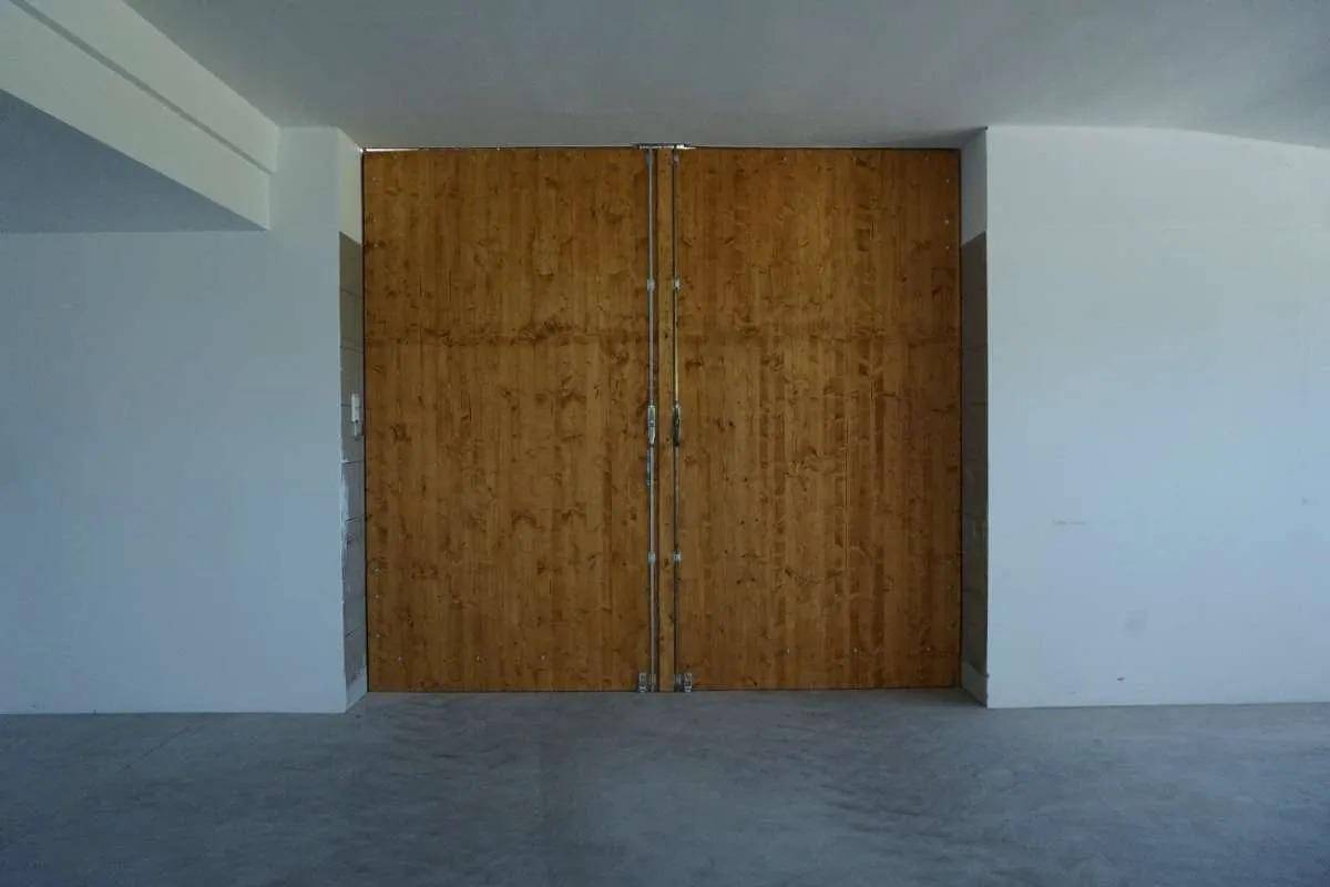 Großer, leerer Innenraum mit Betonboden und weißen Wänden, am Ende befindet sich eine große Holztür.