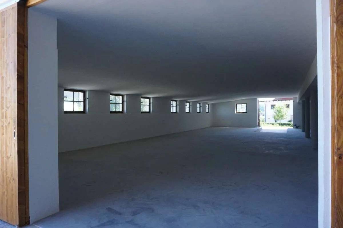 Langer, leerer Innenraum mit Betonboden, weißen Wänden und einer Reihe kleiner Fenster an der Seite.