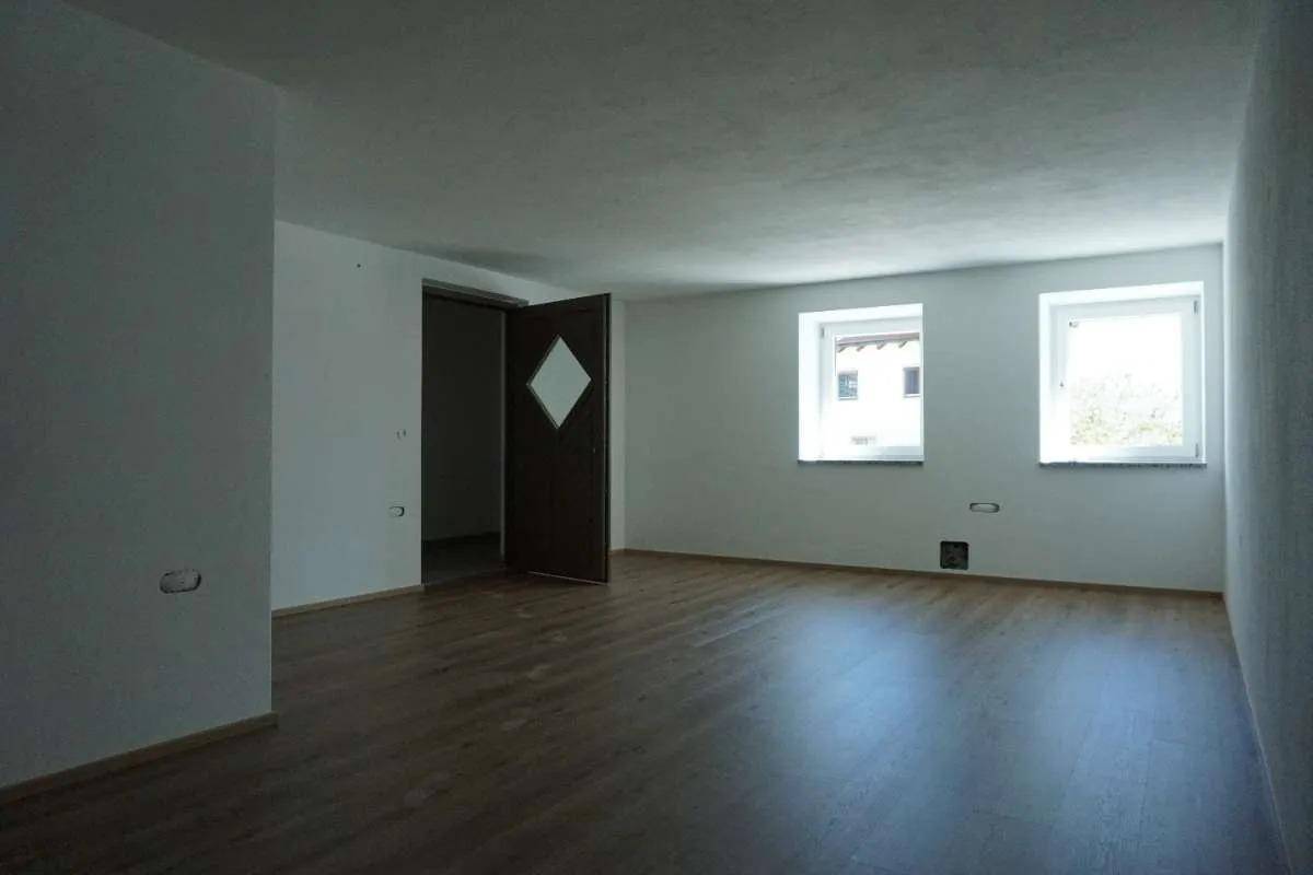 Leerer Raum mit Holzlaminatboden, weißen Wänden und zwei Fenstern, eine Holztür steht offen.